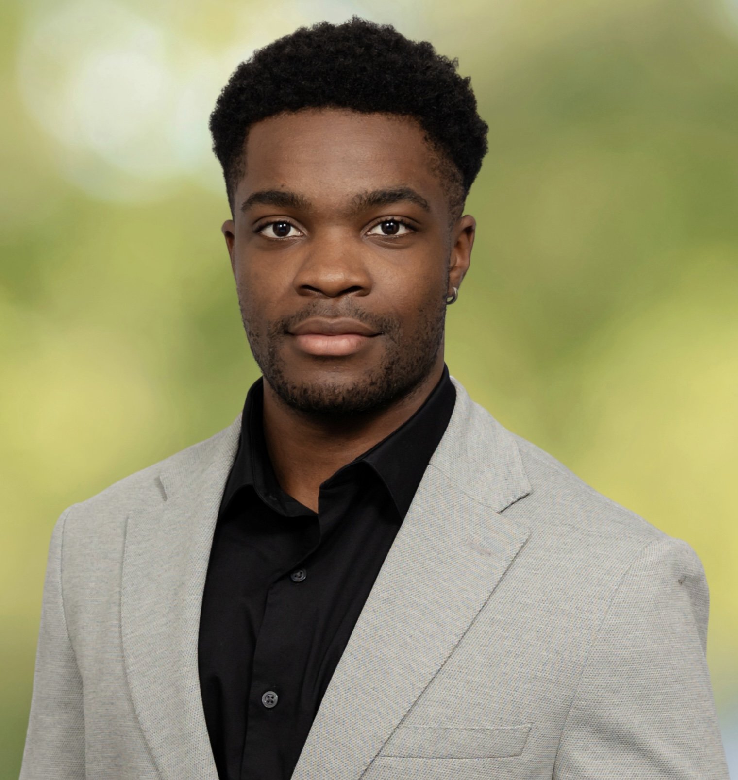 Portrait of a young man with dark curly hair wearing a light gray suit and black shirt, standing against a blurred green and yellow background.