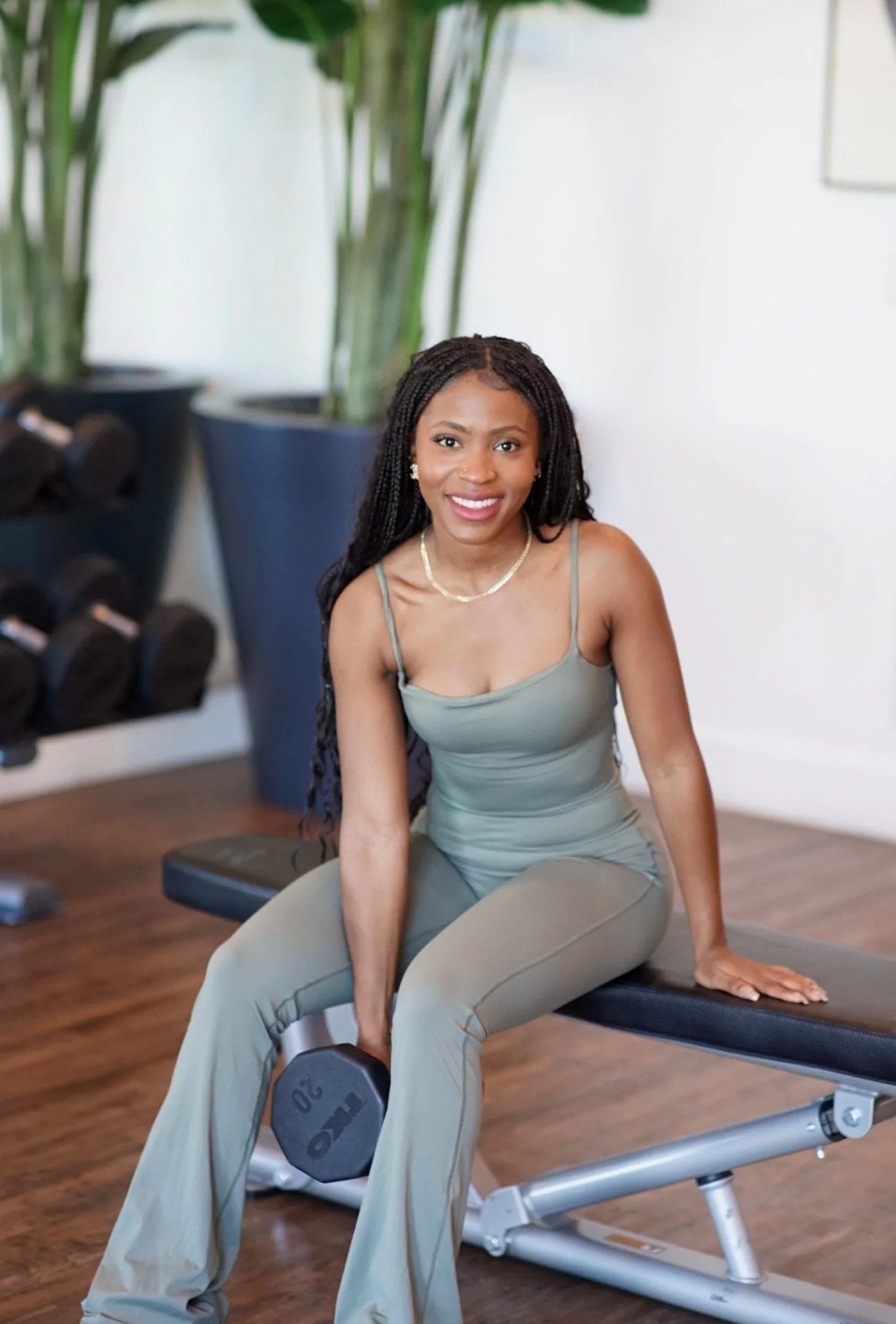A woman with long braided hair, wearing a light green athletic outfit, smiling while sitting on a workout bench in a gym, holding a 20-pound dumbbell.