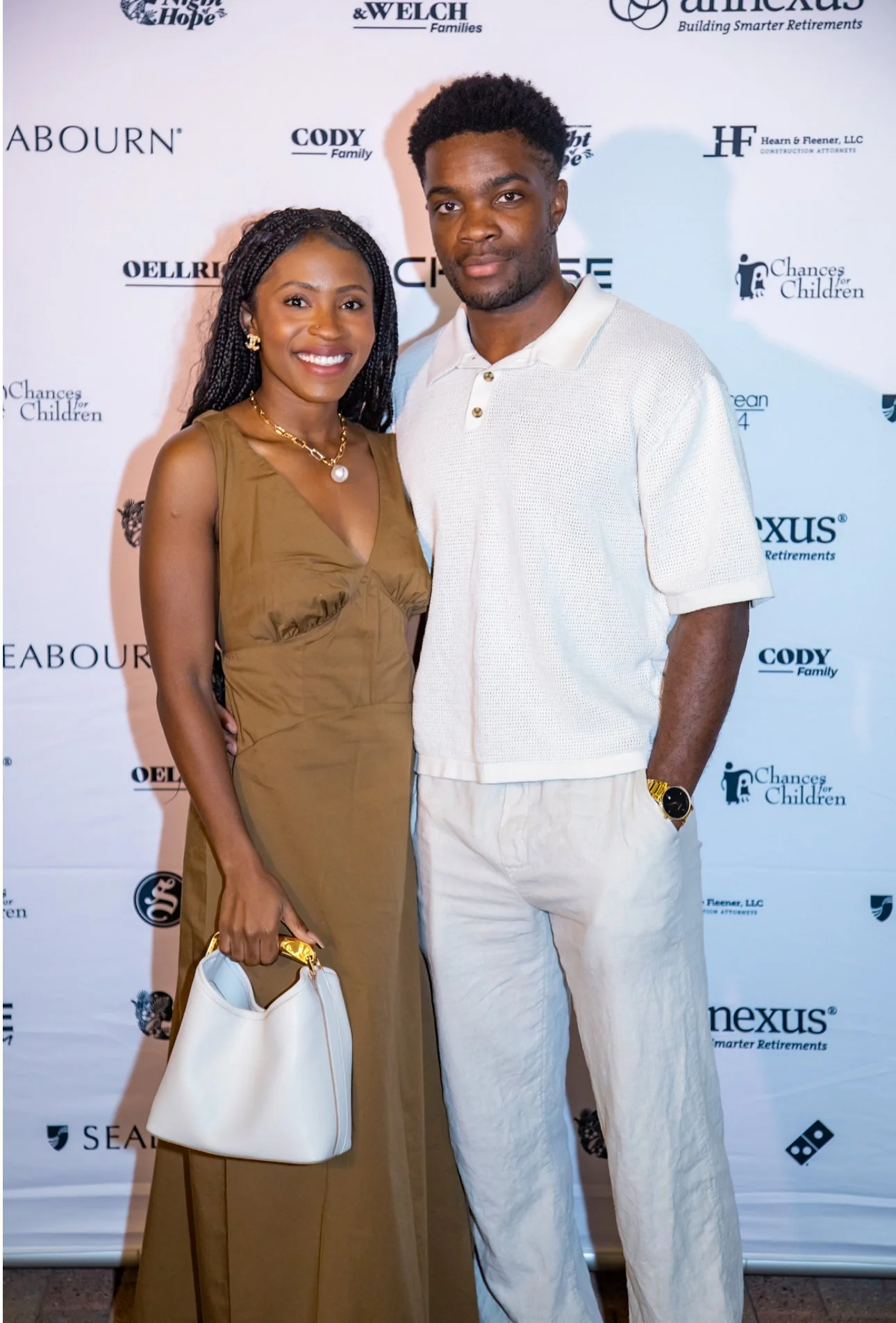 A smiling woman in a tan dress holding a white purse stands next to a man in a white shirt and light-colored pants, both posing in front of a step-and-repeat backdrop with various logos.