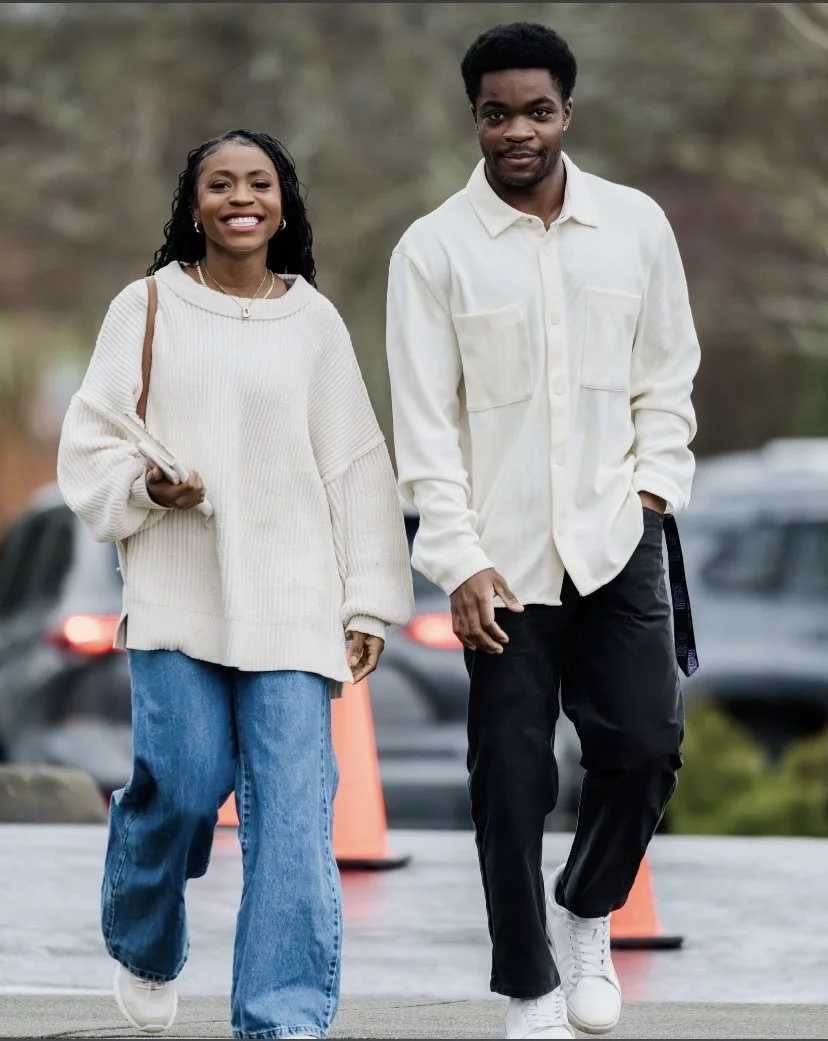 A smiling woman and a man walking outdoors on a cloudy day, dressed in casual clothing, with traffic cones and parked cars in the background.