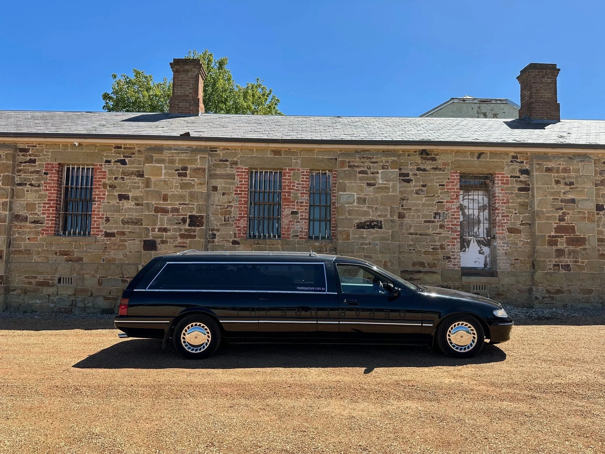 A black hearse with The Departure signage parked in front of a stone building with barred windows, under a clear blue sky.
