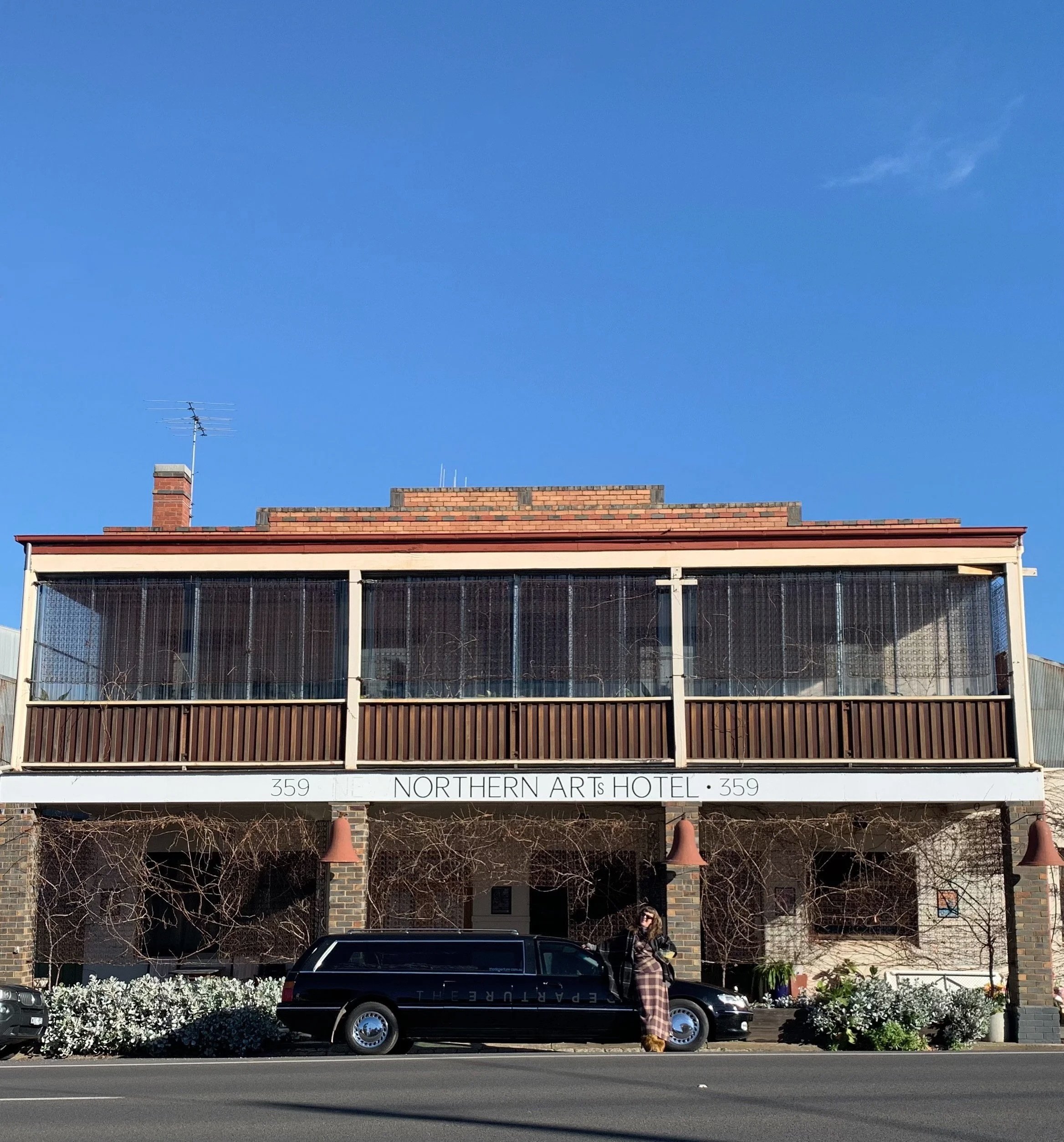 A two-story brick building labeled 'Northern Arts Hotel' with a black hearse parked in front and a woman standing next to it in plaid, autumnal colours, under a clear blue sky.