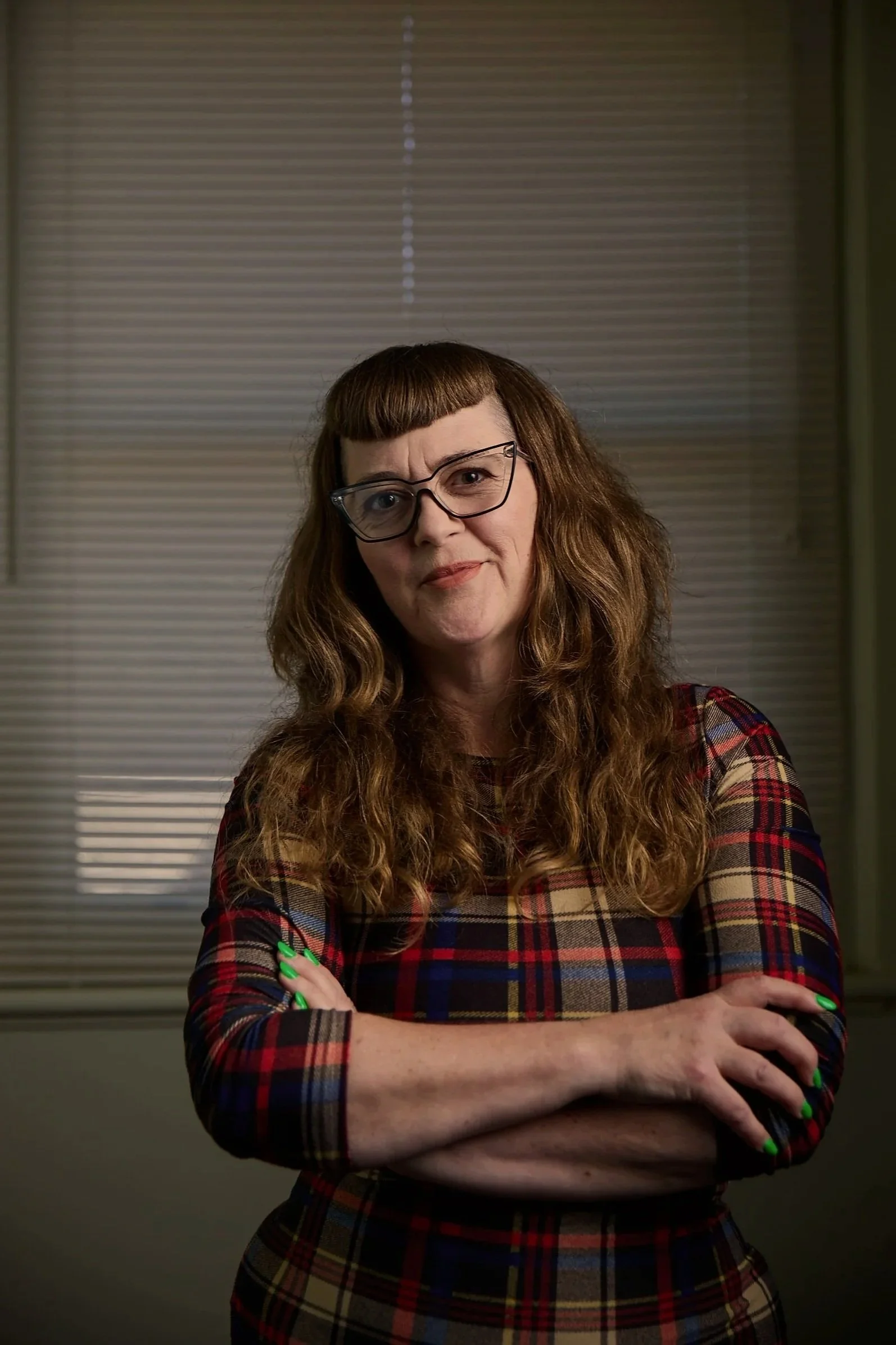 A woman of English and Yenish heritage with wavy dark blonde hair and glasses, wearing a plaid dress, standing with arms crossed with neon green nails, in front of window blinds.