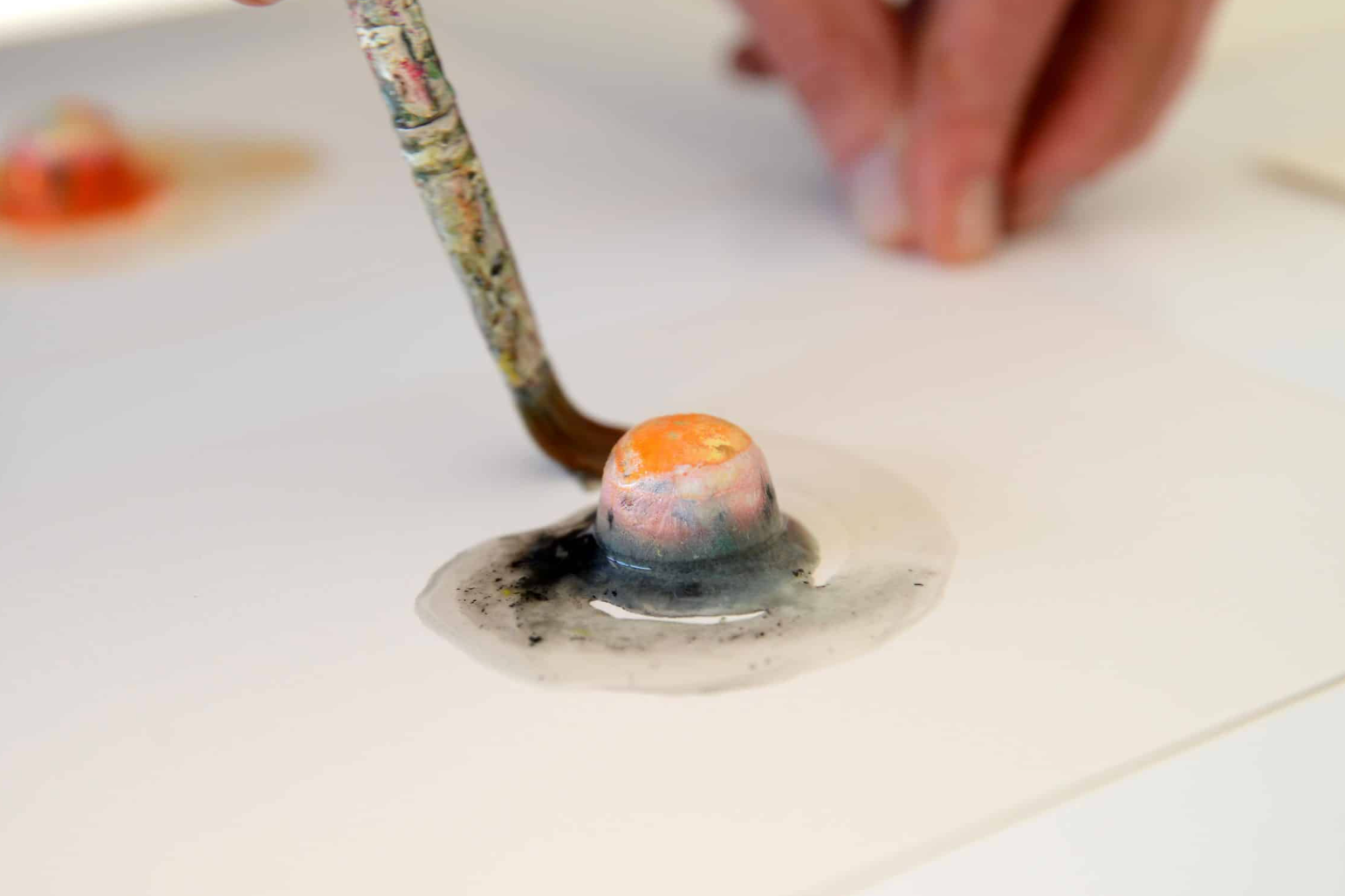 Close-up of a brush mixing dark cremated ashes with orange paint powder and water to create an art work.