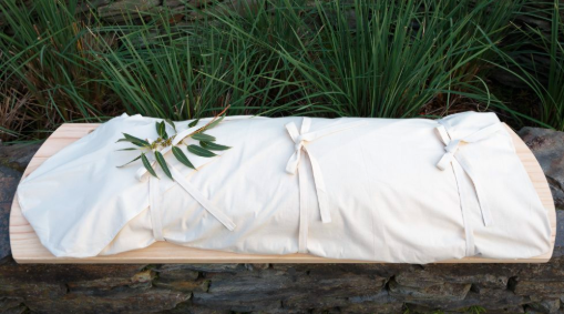 A natural coloured bamboo shroud wrapped around a body sits on a wooden shroud carrier. The carrier and shroud sits amongst grass.
