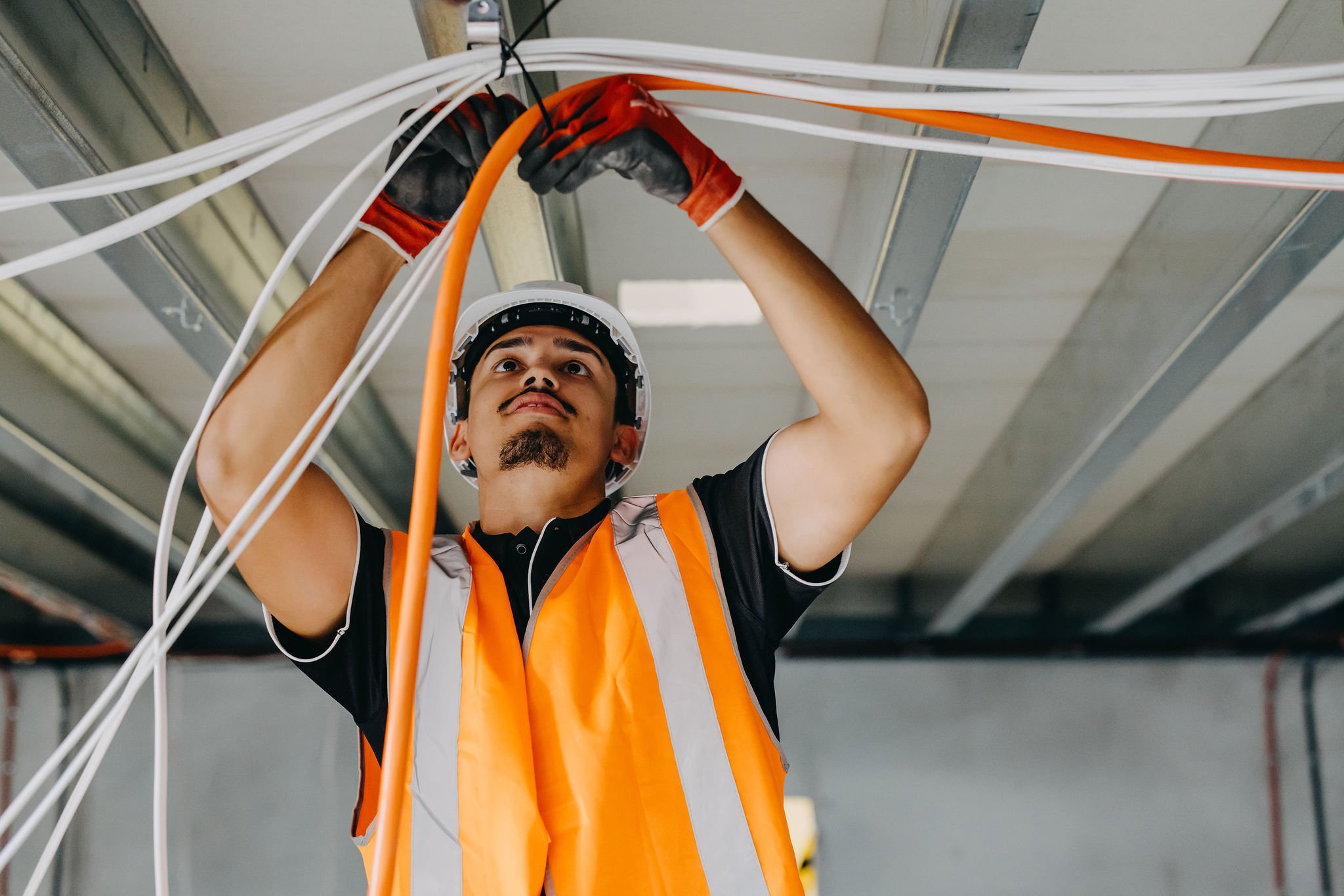 A technician wearing a safety helmet, orange safety vest, and gloves working on electrical wiring in an industrial or commercial setting.