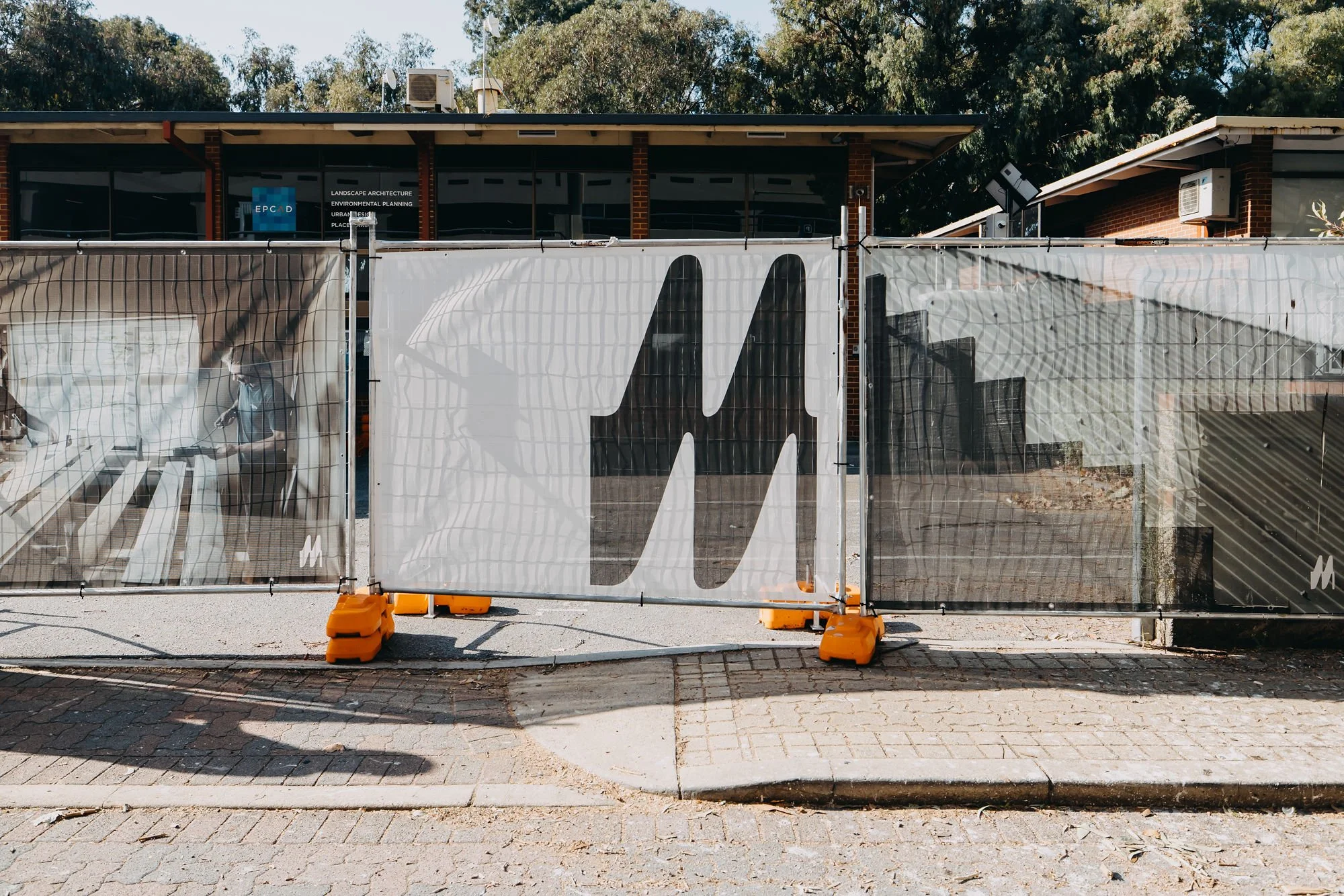 Construction site with a temporary fence and a building in the background. The fence has a large graphic design and is secured with orange sandbags. The building is made of brick with windows and is partially obscured by trees.