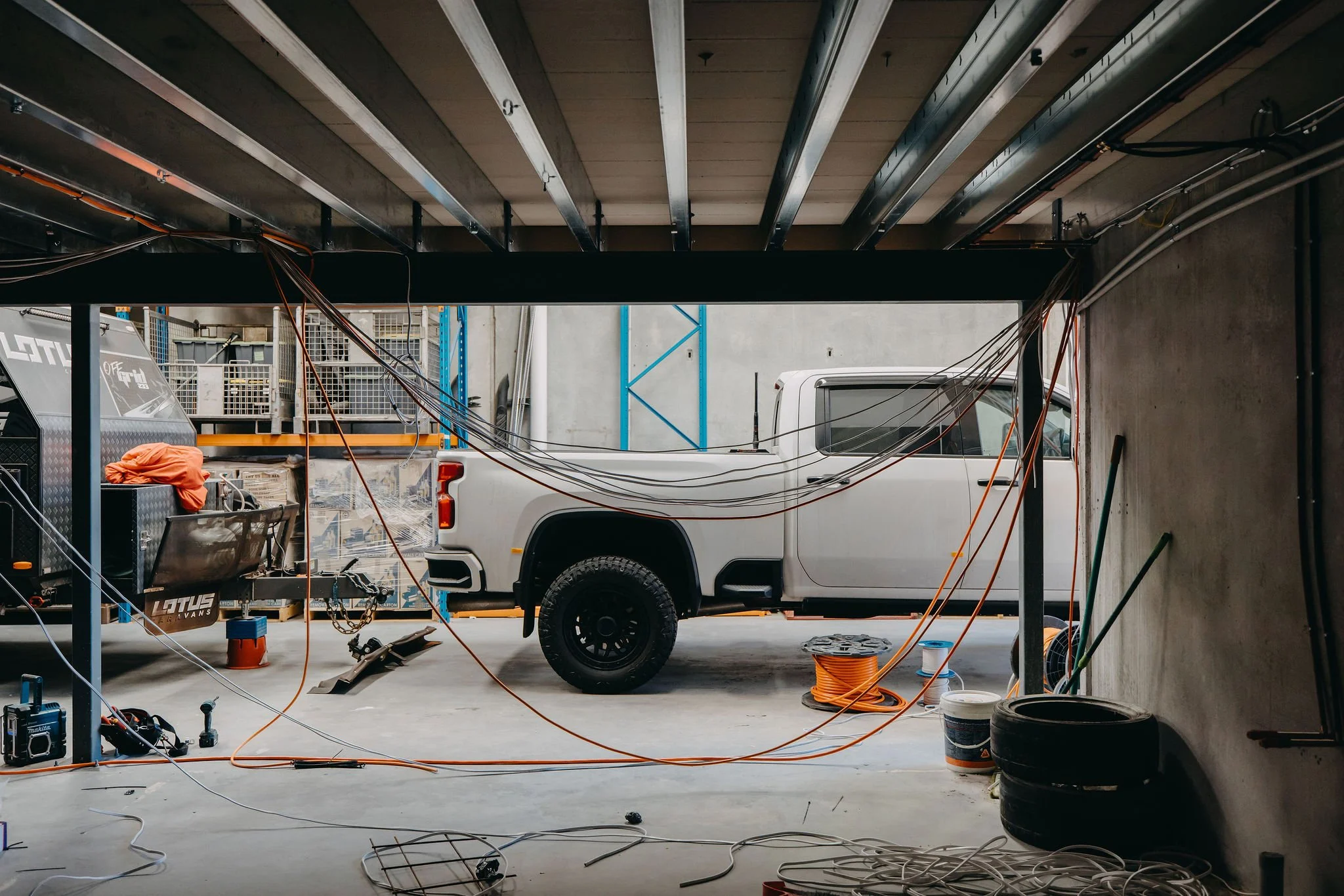 Construction or workshop scene with a white pickup truck, electric cables, tools, and equipment. The ceiling is exposed with metal beams, and there is a storage rack and other construction materials in the background.