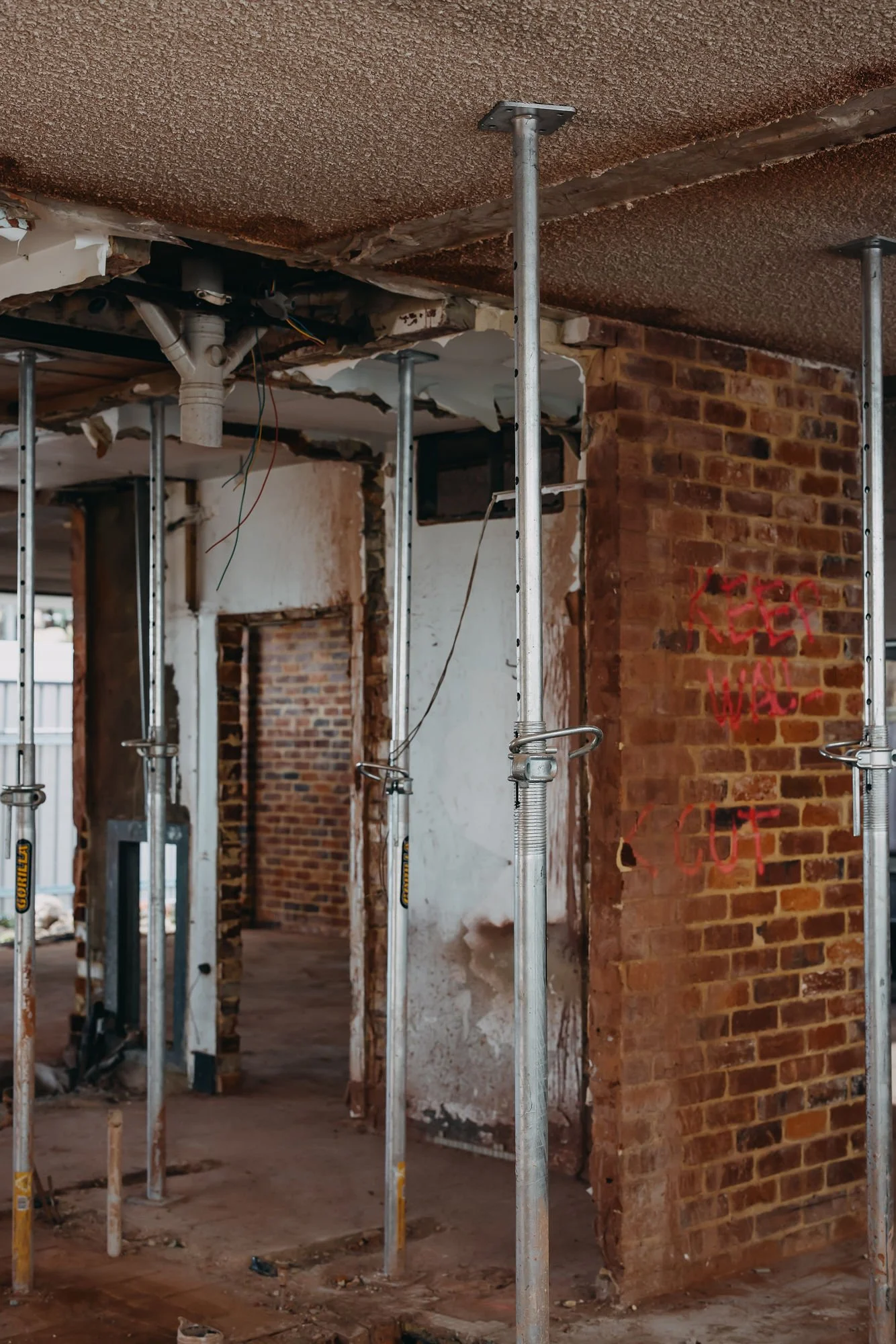 Interior of a building under renovation with exposed brick walls, metal scaffolding poles, and ceiling that is partially torn down.