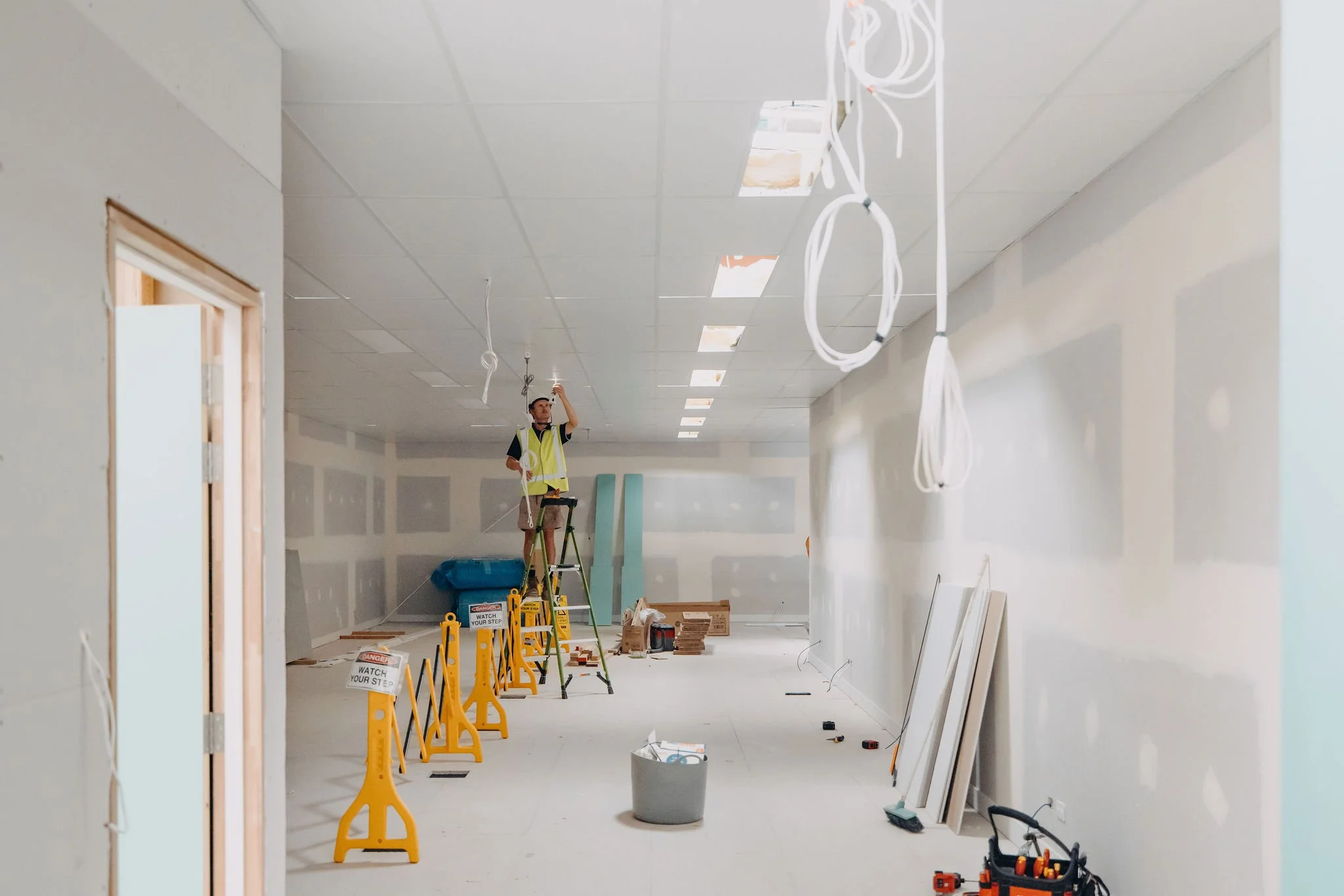 Construction worker standing on ladder installing or inspecting ceiling panels in a building under renovation. Wires hang from the ceiling, and safety signs are placed on the floor.