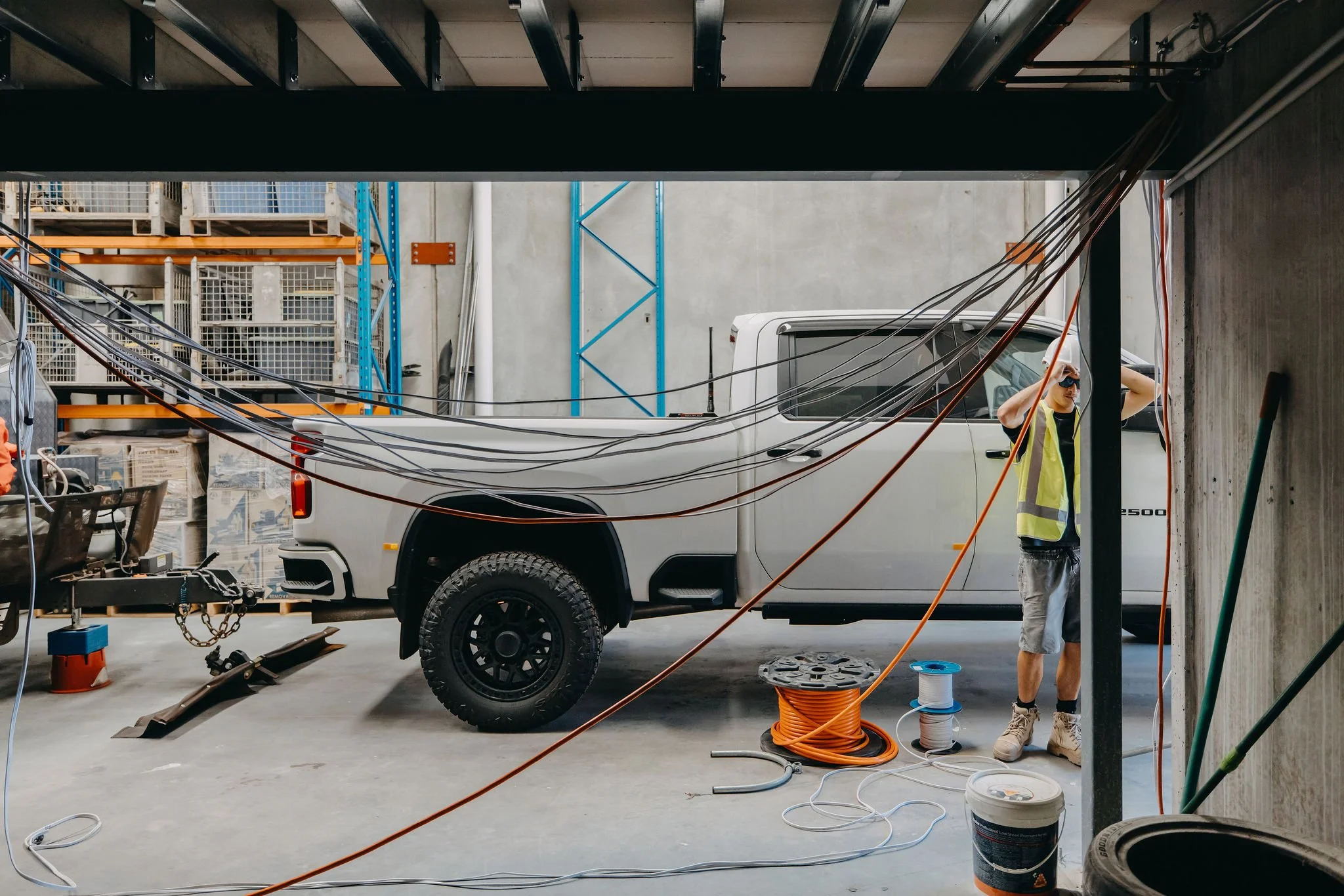 An indoor construction site with a white pickup truck and a person wearing a yellow safety vest and cap, standing next to cables and construction materials.