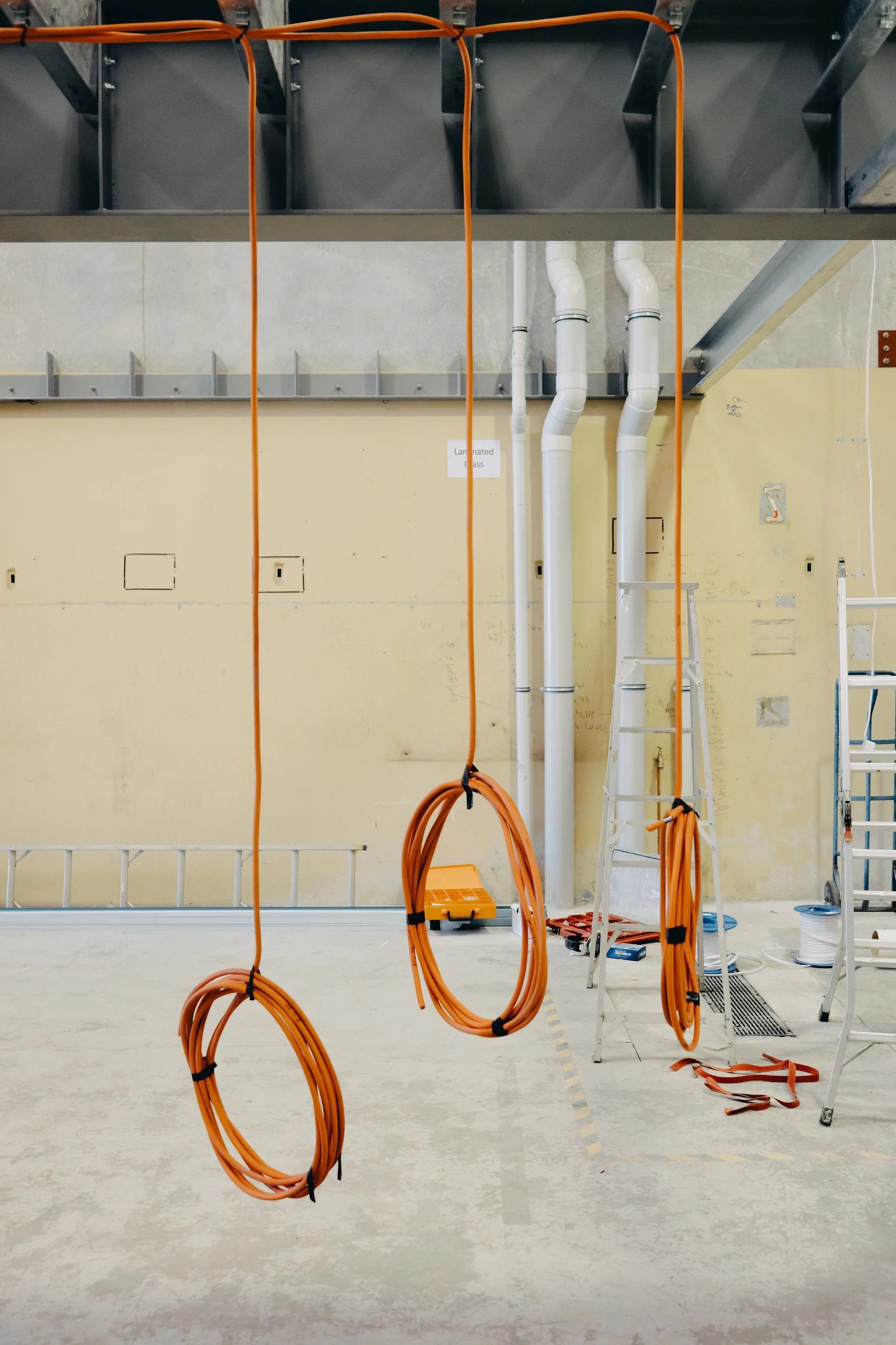 Construction site with orange extension cords hanging, ladders, and construction materials. A sign reading 'Laminated Glass' is on the wall. Pipes run vertically, a ladder is beside the pipes, and various tools and materials are scattered on the floo