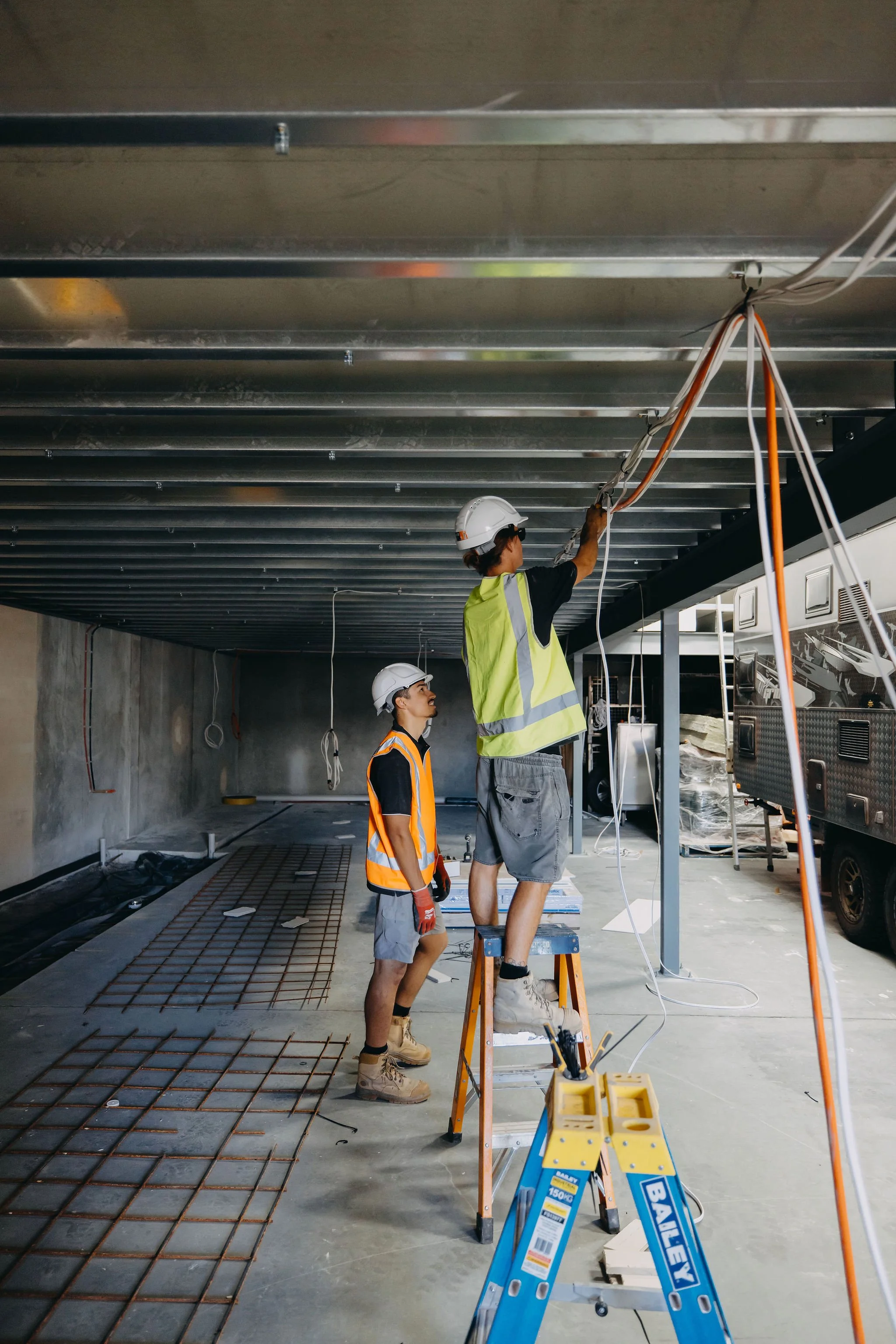 Construction workers wearing safety gear installing electrical wiring on a ceiling inside a building.