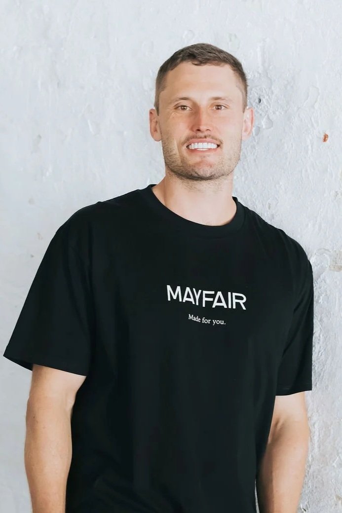 A smiling man with short brown hair and light skin, wearing a black T-shirt with the words "MAYFAIR Made for you." printed on it, standing against a white textured wall.