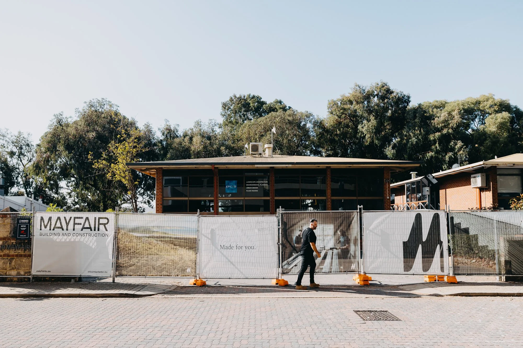Construction site fence with banners, one reading 'MAYFAIR Building and Construction,' in front of a building under renovation, with trees and a clear sky in the background, and a person walking past.