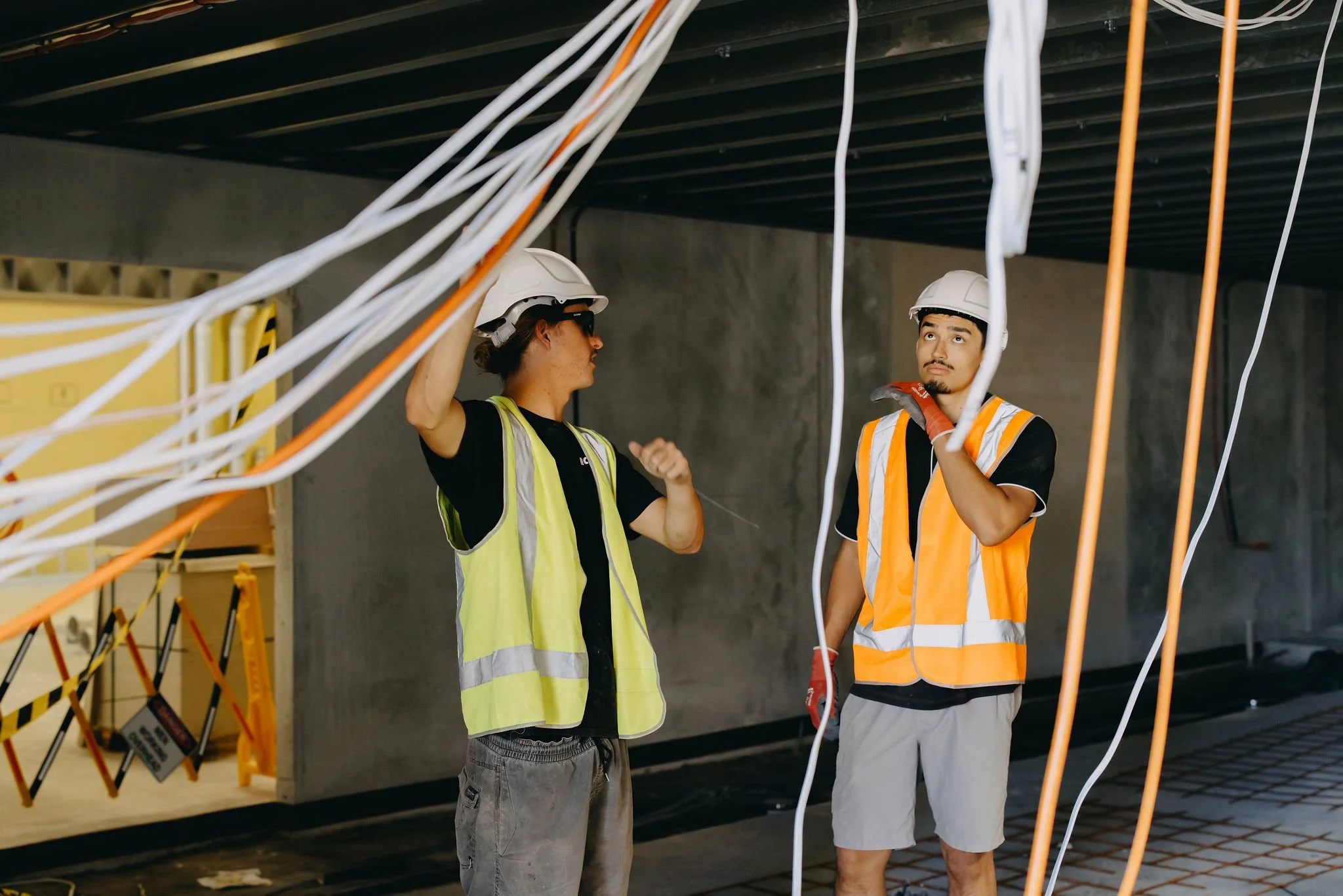 Two construction workers, one in a yellow vest and the other in an orange vest, wearing white helmets, are working with electrical cables in an unfinished building.