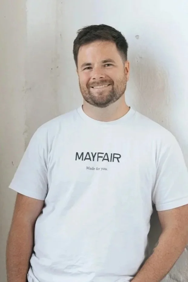 A man with dark hair and a beard, smiling, wearing a white T-shirt that says "MAYFAIR Made for you" in front of a light-colored wall.
