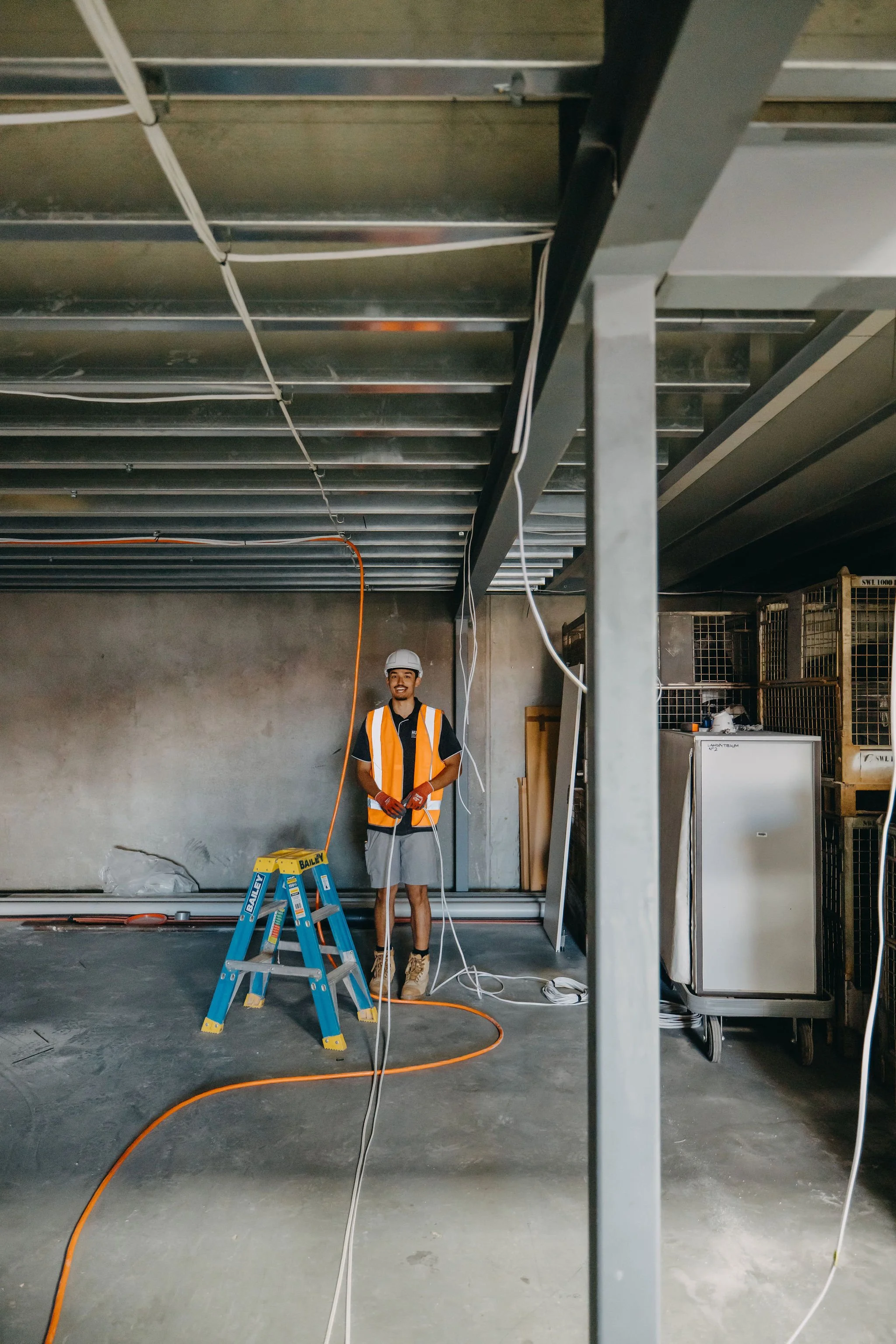 A construction worker in a white helmet and orange safety vest stands in a building under construction, smiling at the camera. He is holding an extension cord, with a ladder nearby, and various wires and construction materials are visible around him.