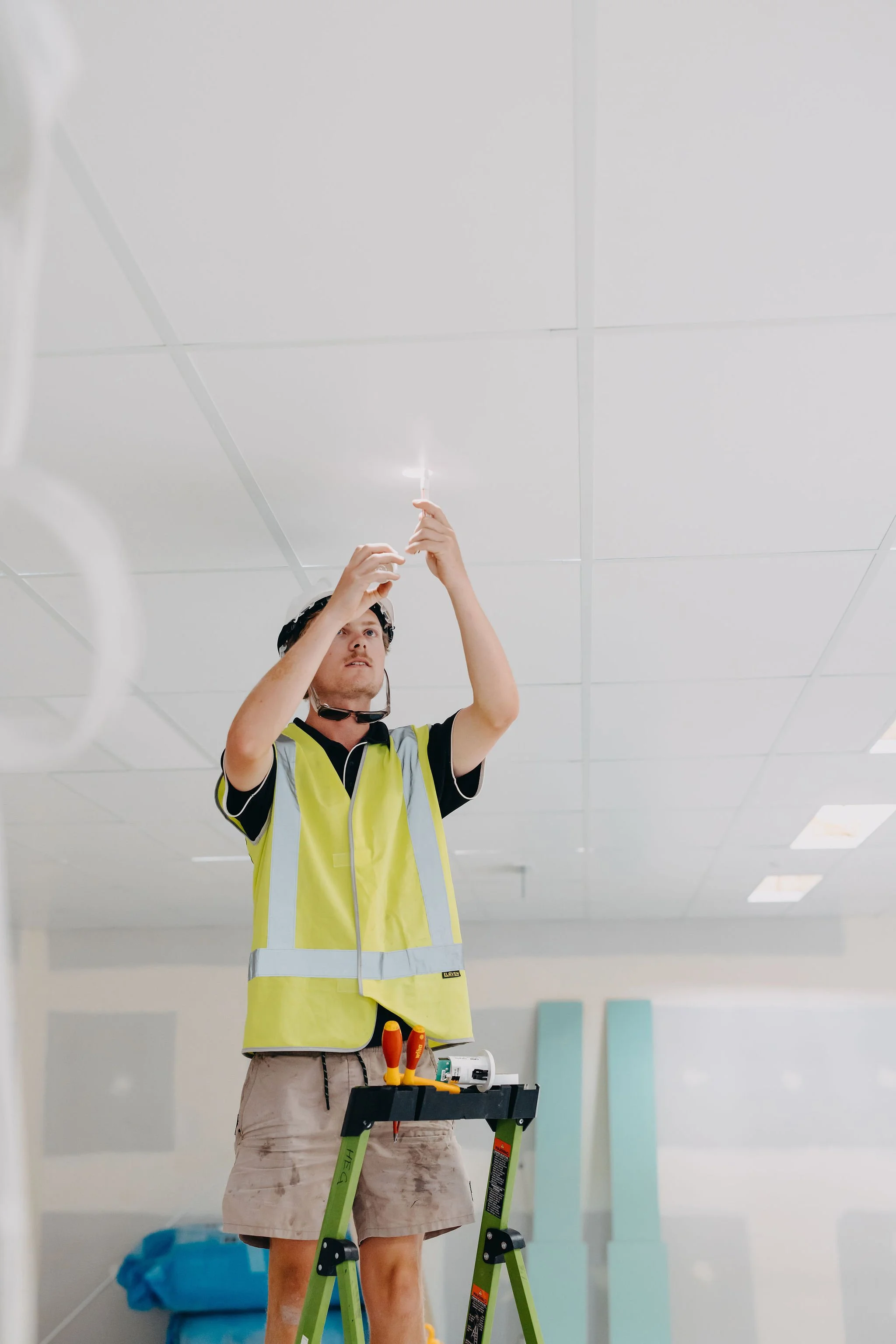 Worker standing on a green ladder, wearing safety goggles and a yellow safety vest, installing or inspecting ceiling tiles in a large, well-lit interior space.