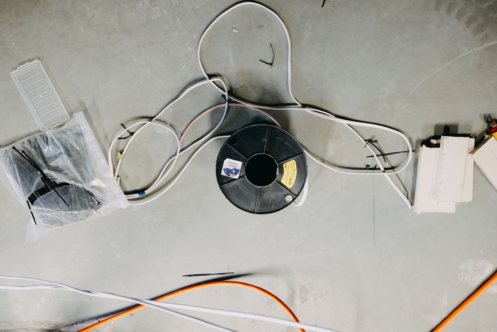 Various electrical wires and cords on a concrete floor, including a black spooled wire, a plastic bag with electrical zip ties, and a small white plastic component.
