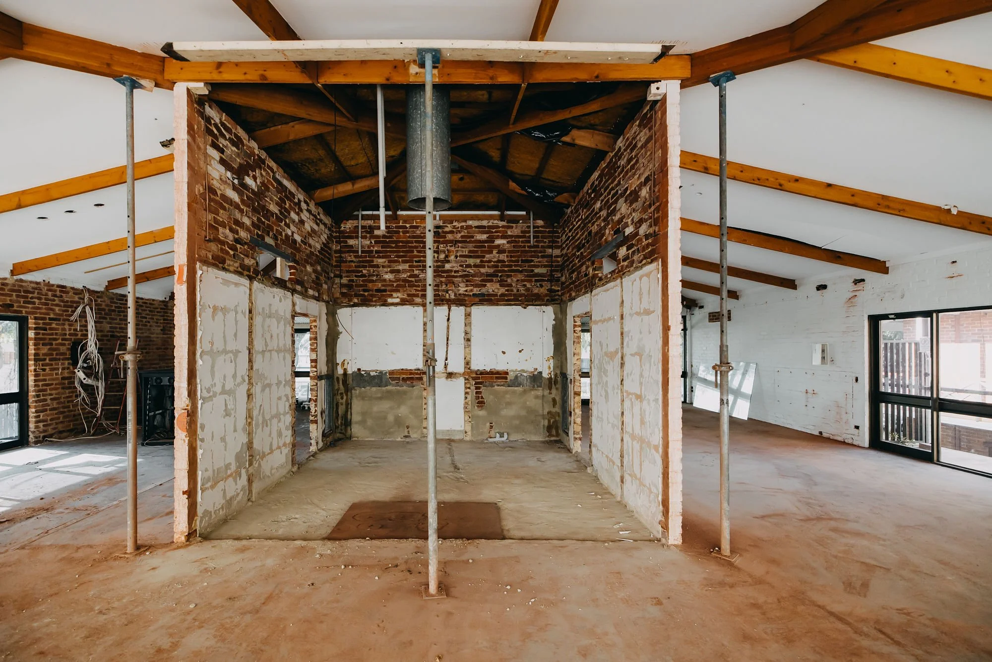 Interior view of a house under construction with exposed brick walls, wooden ceiling beams, and support poles, unfinished floors, and construction materials.