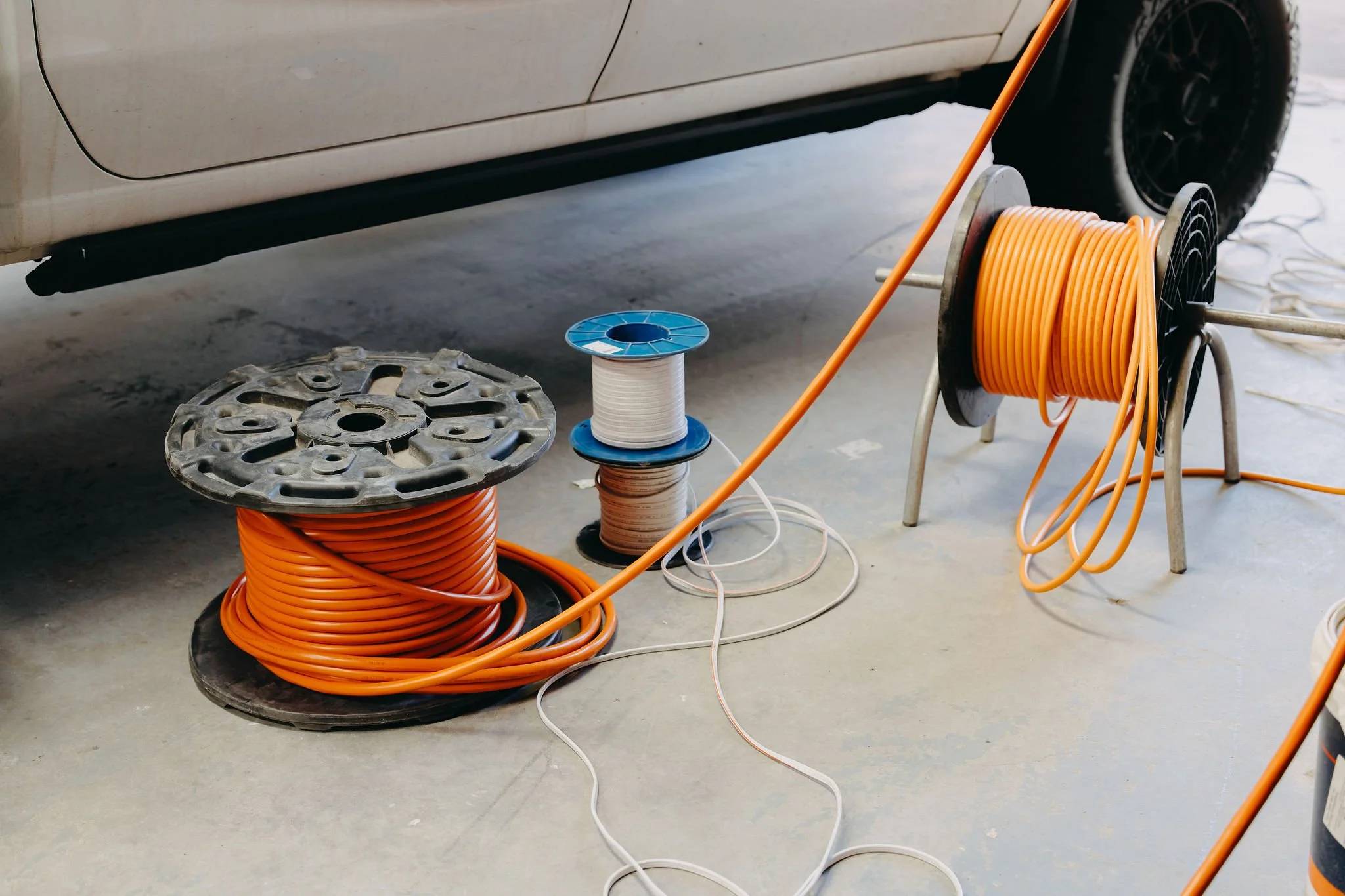 Orange extension cords and spools of wiring on the garage floor near a white vehicle.