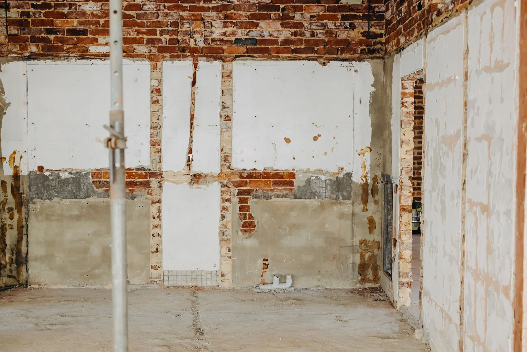 Interior of a building under construction with exposed brick walls, drywall framing, and a concrete floor. A metal pole is in the foreground, and electrical wiring is visible near the ceiling.