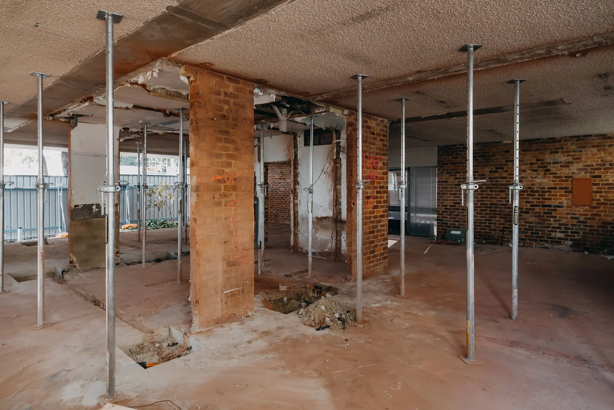 Interior of a building under renovation with brick columns, exposed ceiling, construction supports, and holes in the floor.