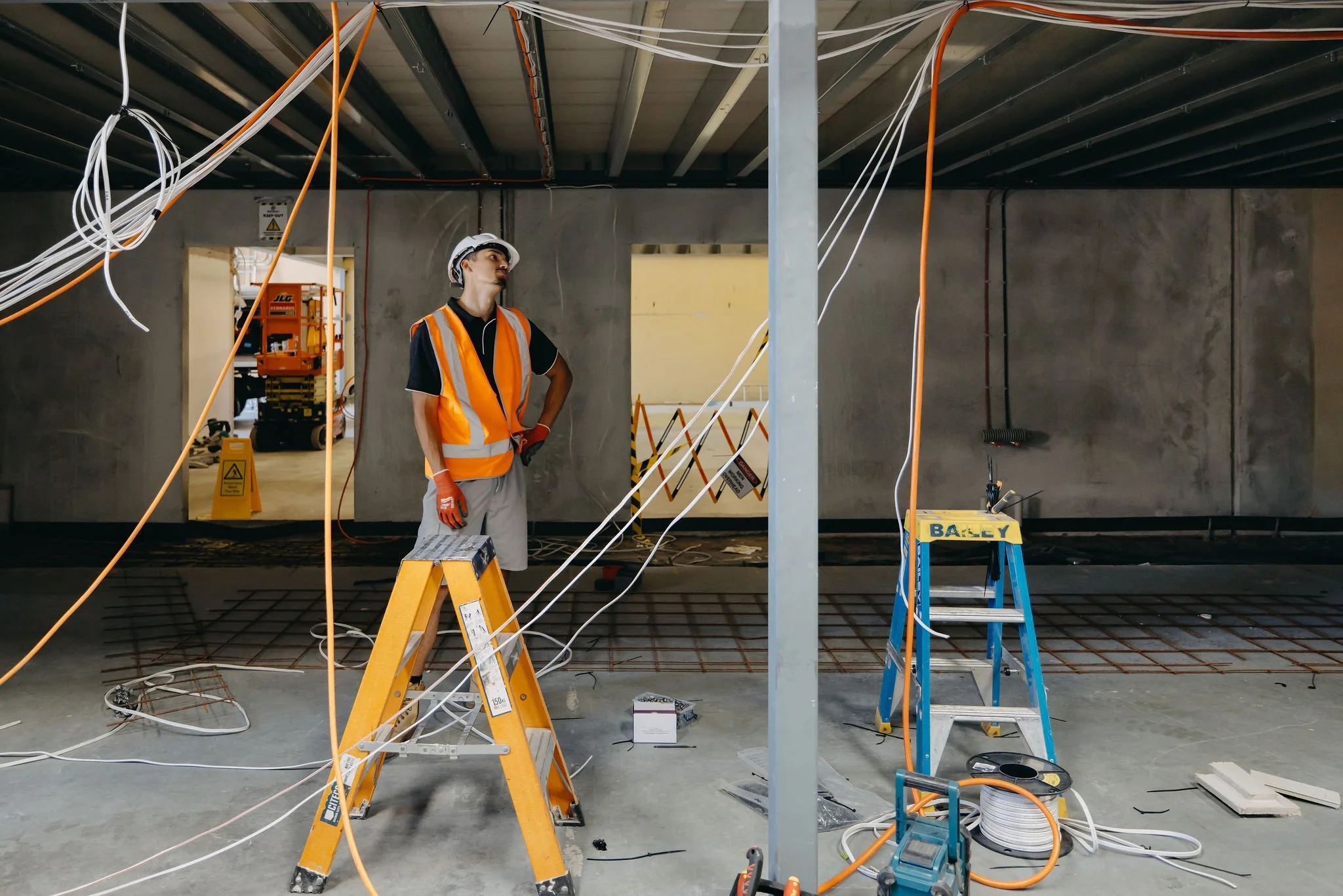 A construction worker in an orange safety vest and white hard hat stands inside a building under construction surrounded by hanging wires, tools, and scaffolding.