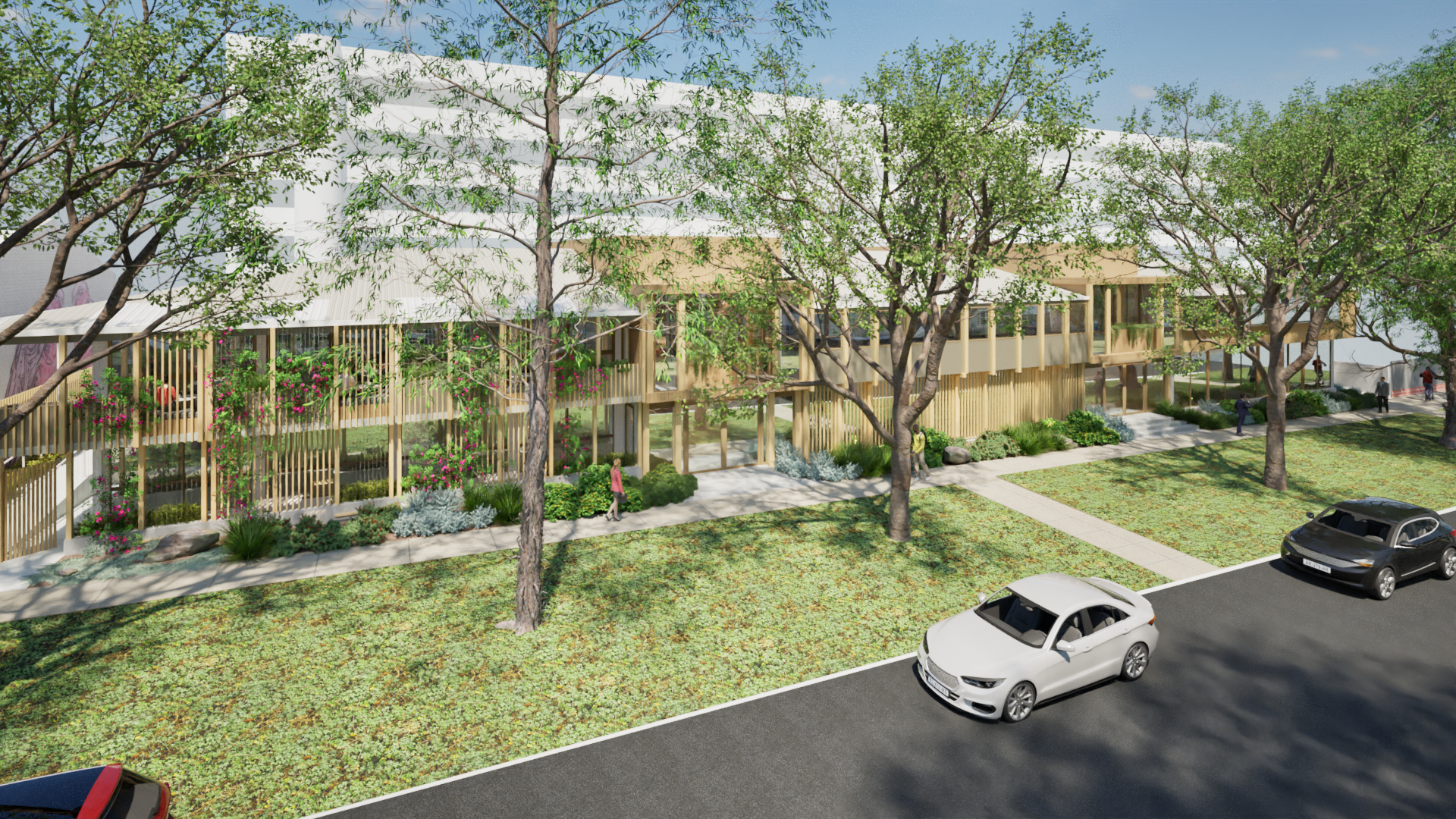 A modern multi-story building surrounded by trees and landscaped greenery, with a sidewalk and several parked cars in the foreground. People are walking around the area.