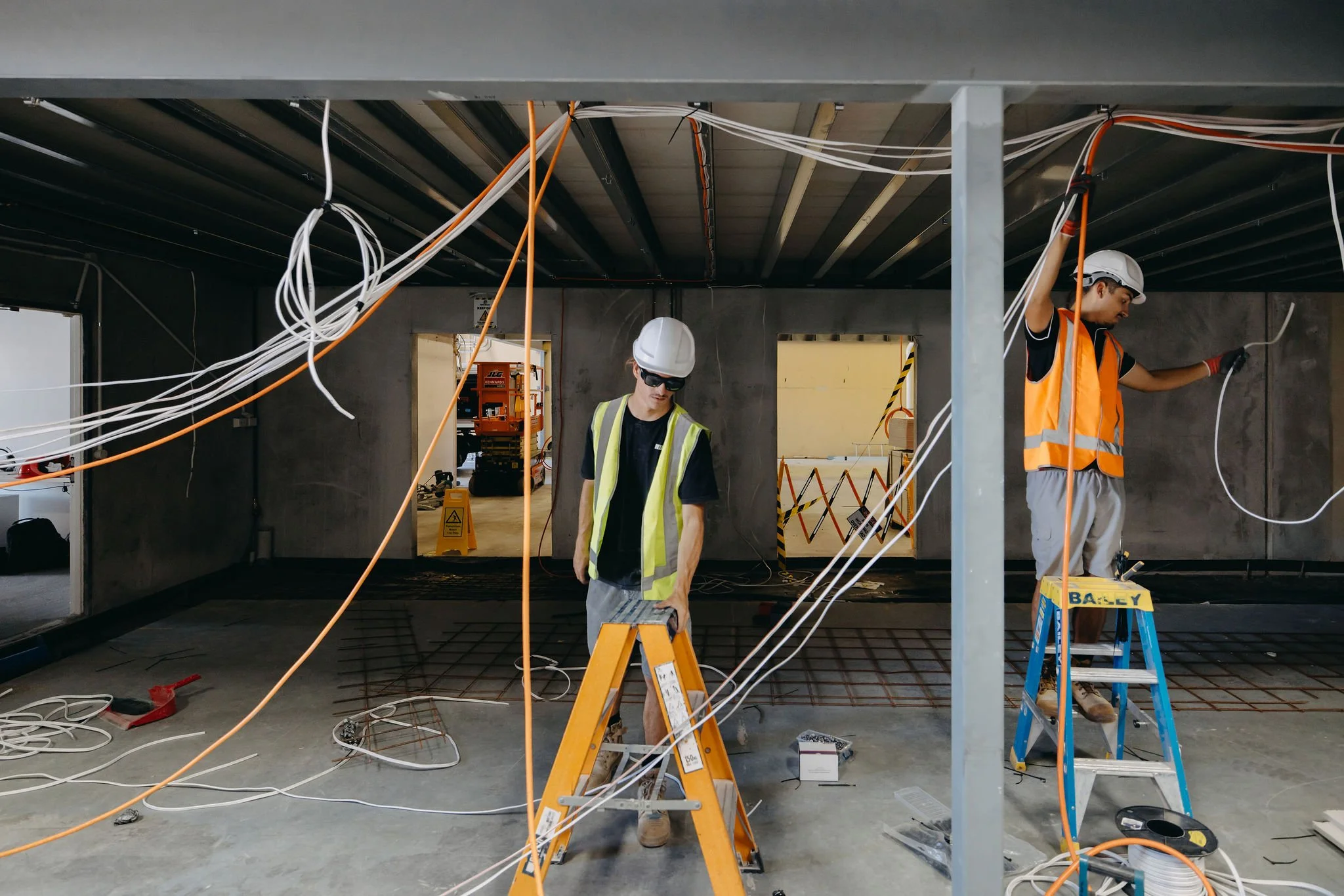 Two construction workers wearing hard hats and safety vests working on wiring inside a building under construction. One is standing at a ladder and the other is holding wiring, with construction tools and equipment around.