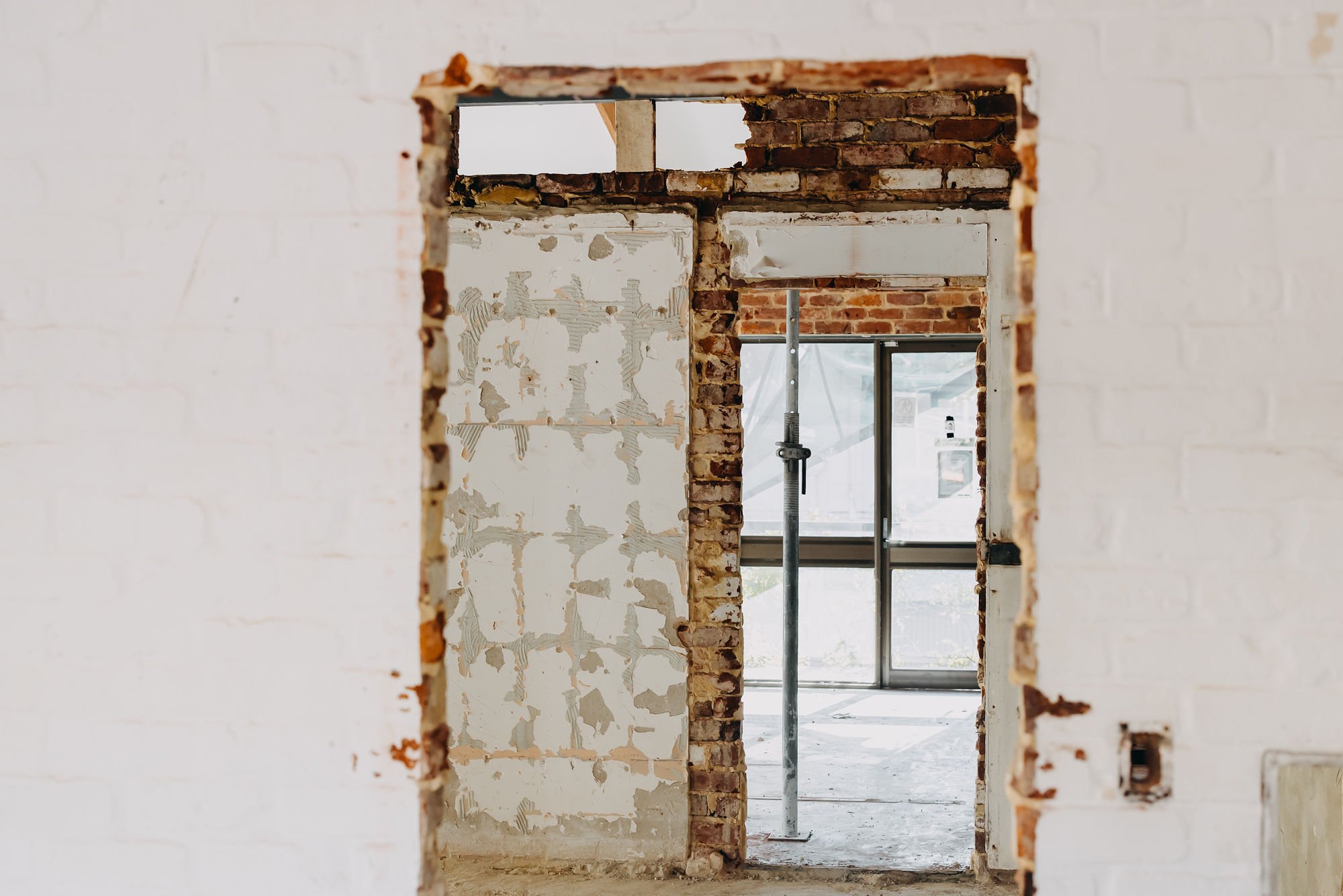 View through a partially demolished interior wall revealing brick and drywall construction, with a window in the background and construction equipment inside.