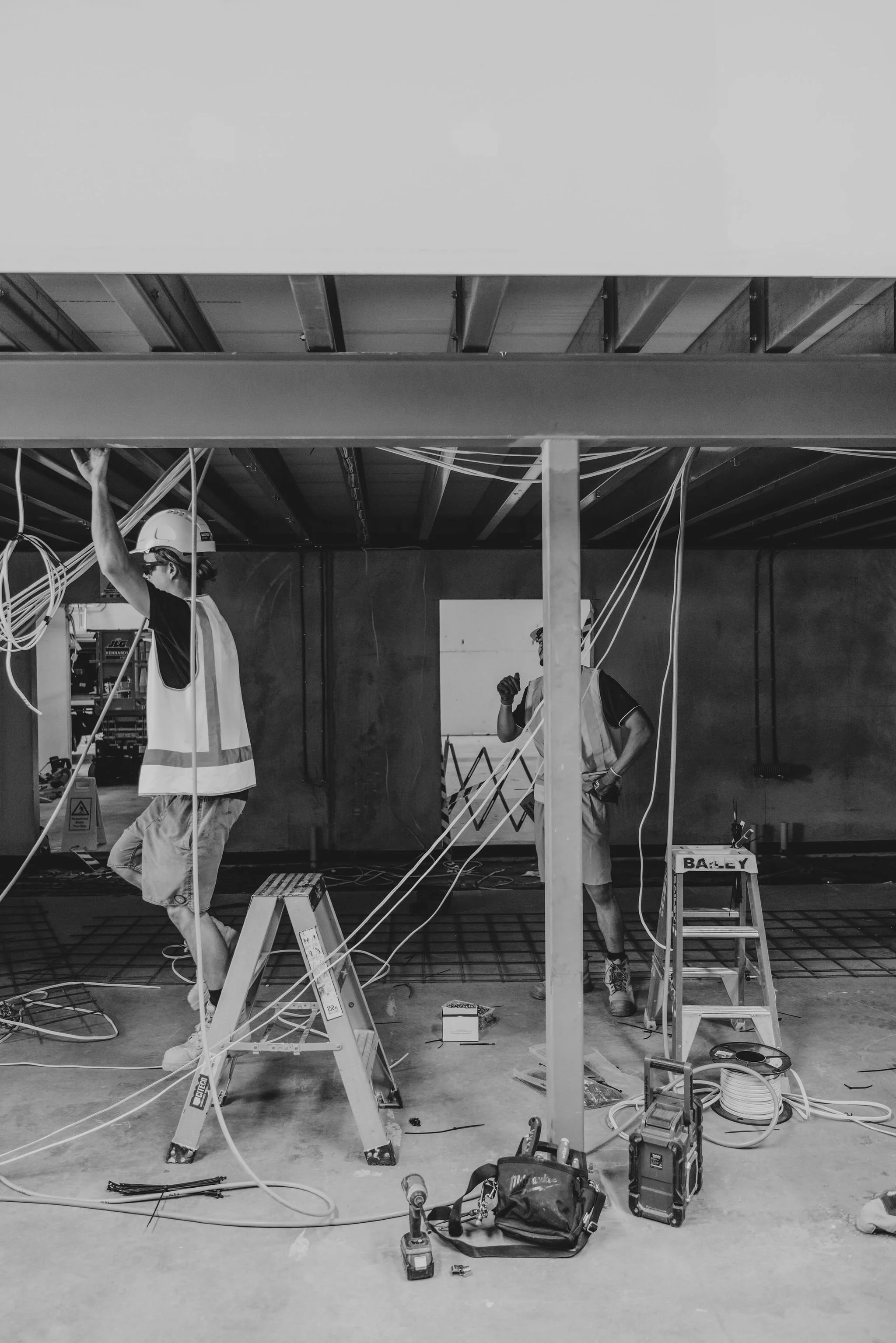 Construction workers wearing safety helmets and vests working inside a building under construction, with tools, ladders, and electrical wiring visible.