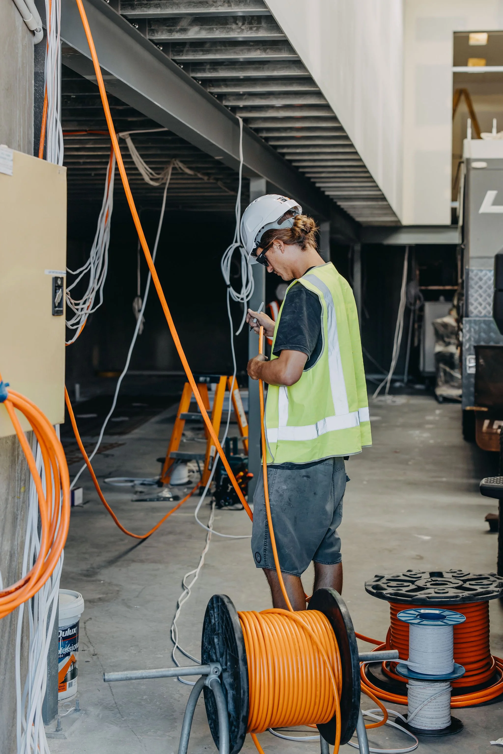 A construction worker with a safety helmet and reflective vest working with electrical wiring in an industrial or construction setting. Various cables, extension cords, and electrical equipment are visible.