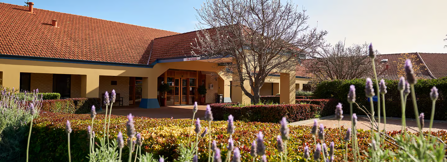Facade of a residential building with a red-tiled roof, surrounded by landscaped garden with flowering plants and trees.