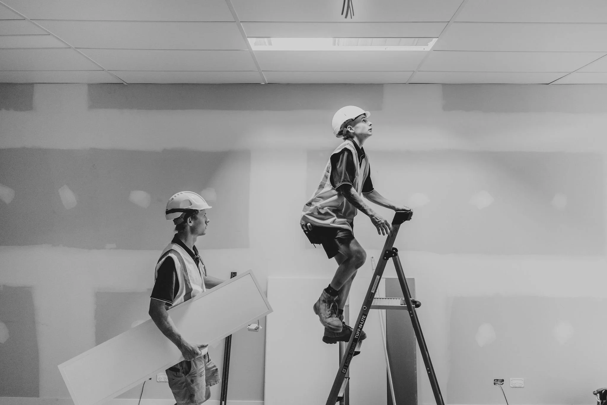 A black and white photo of three construction workers in safety helmets. One worker stands on an elevated platform or ladder, looking upward, and is holding a tool or device. Another worker walks while carrying a large rectangular object, and a third