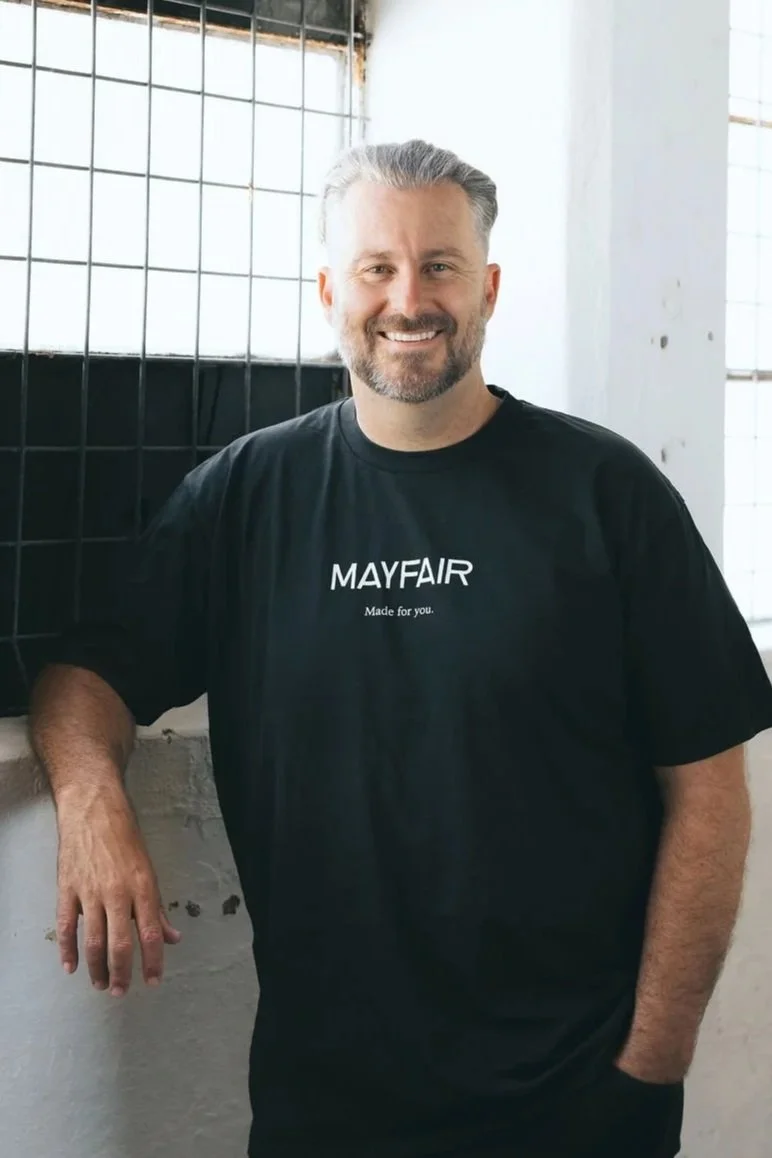 A man with gray hair and a gray beard smiling, wearing a black T-shirt with 'MAYFAIR Made for you' written on it, standing inside a bright industrial-style space with white walls and a wire grid window.
