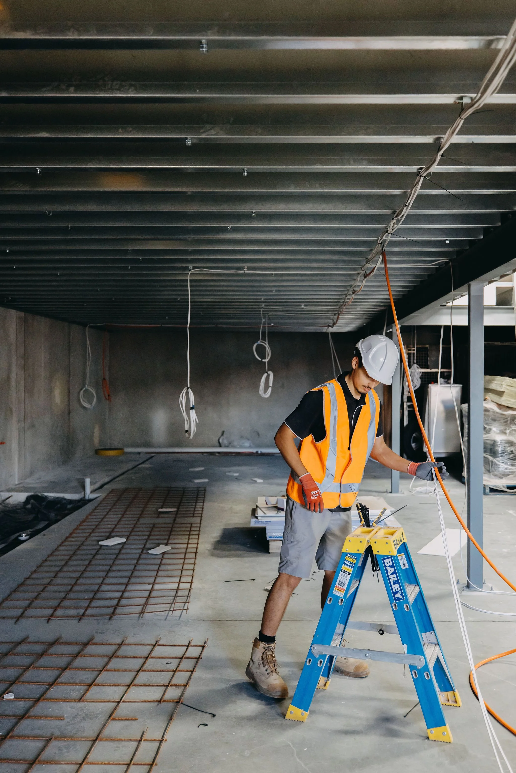 Construction worker in a safety vest and helmet standing on a ladder on a construction site. The ceiling is exposed with metal beams and hanging wires. Work materials and tools are visible on the floor.