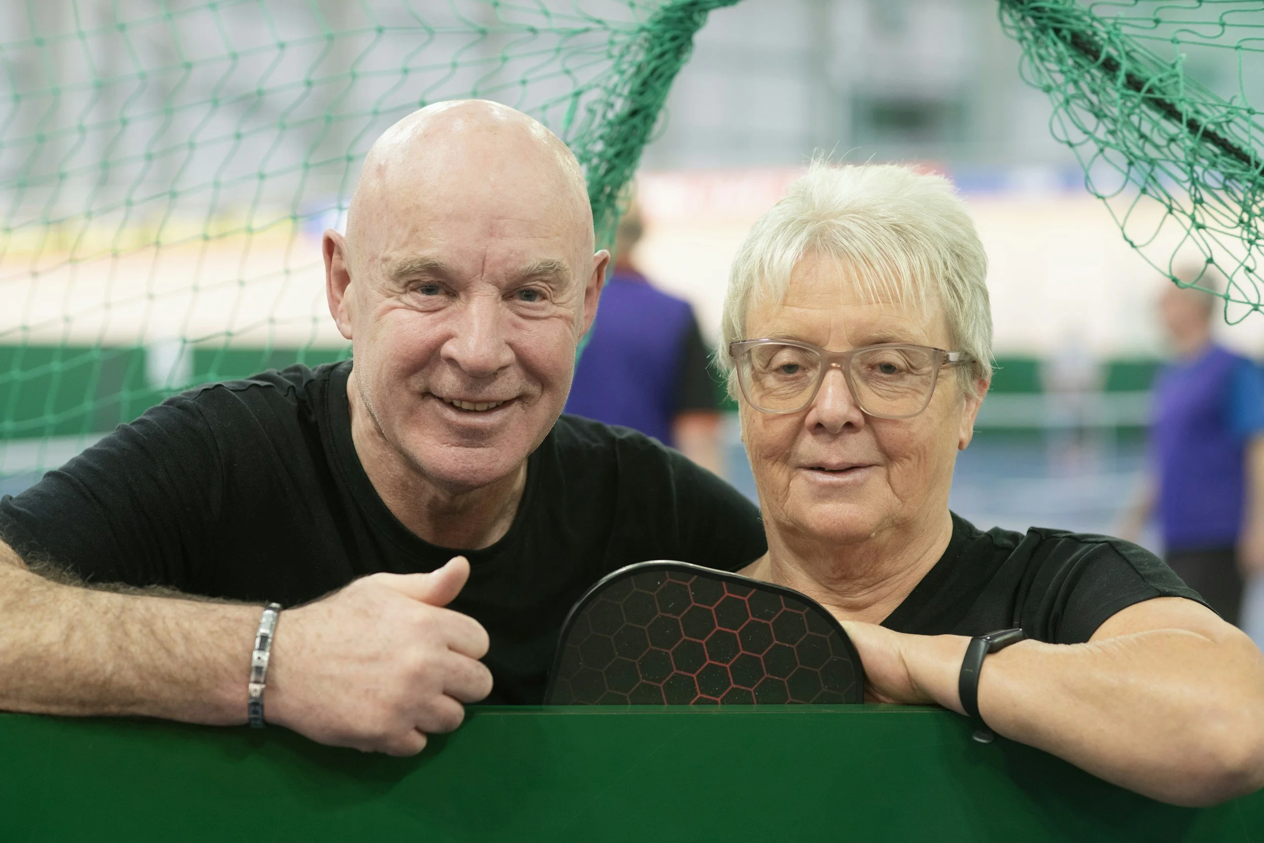 A smiling man and woman in a sports setting, sitting behind a green table with a black paddle, with a sports net and people in the background.