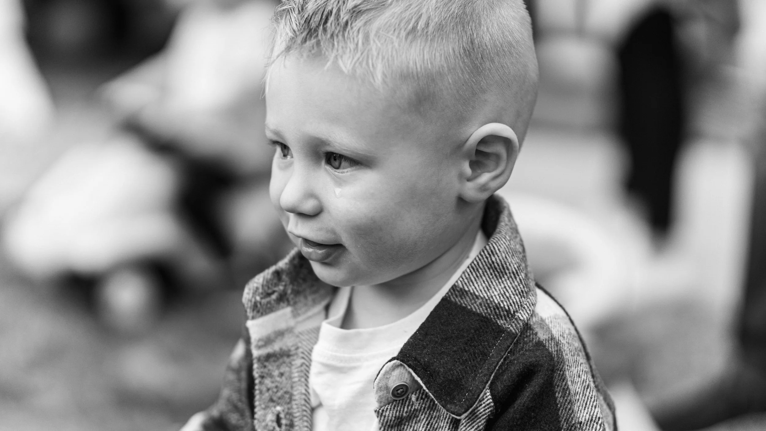 Black and white image of a young child with short hair and a plaid jacket, looking to the side with a tear on their cheek.