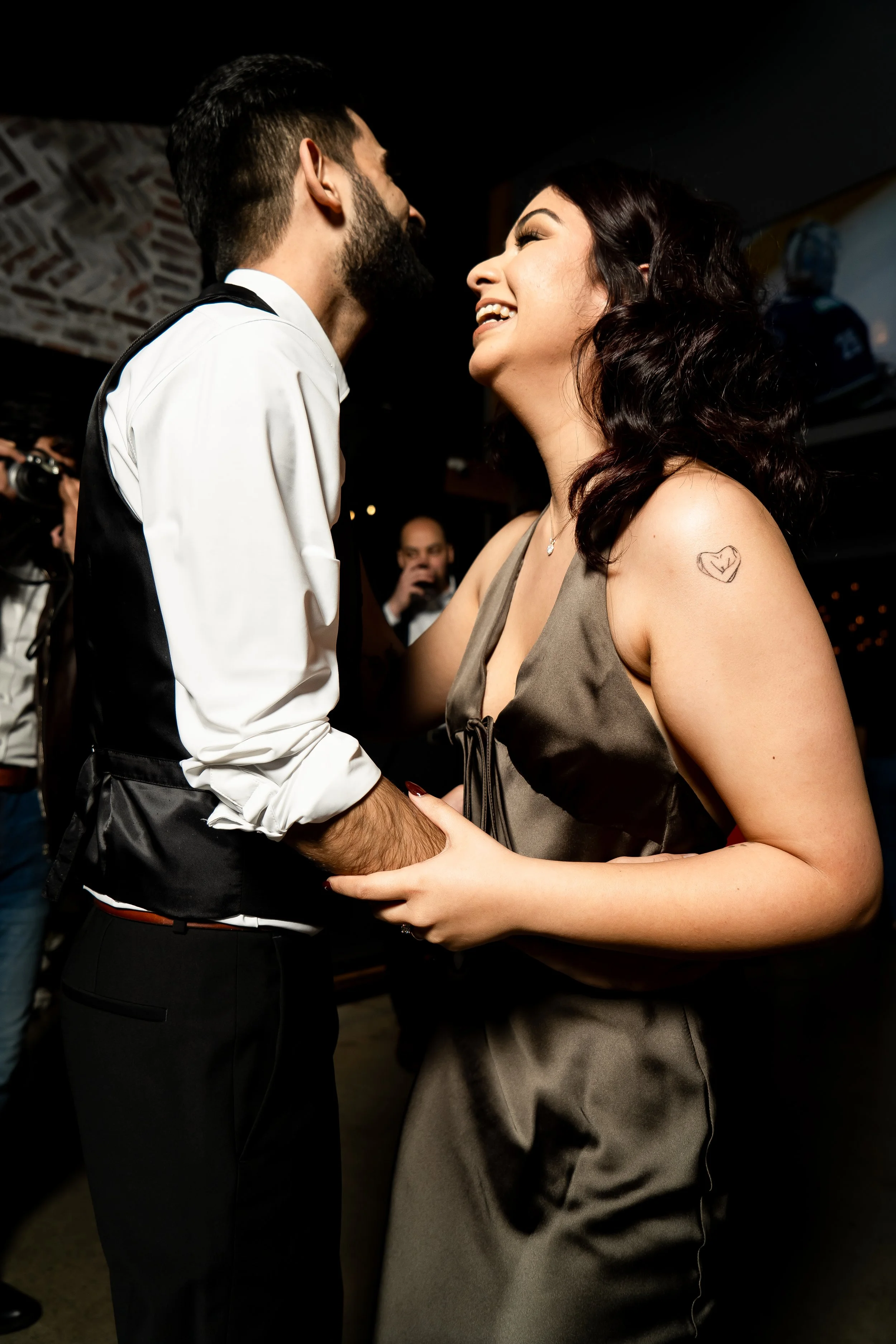A man and woman dancing at an event, looking happy. The woman is wearing a sleeveless dress and has a simple heart tattoo on her shoulder.