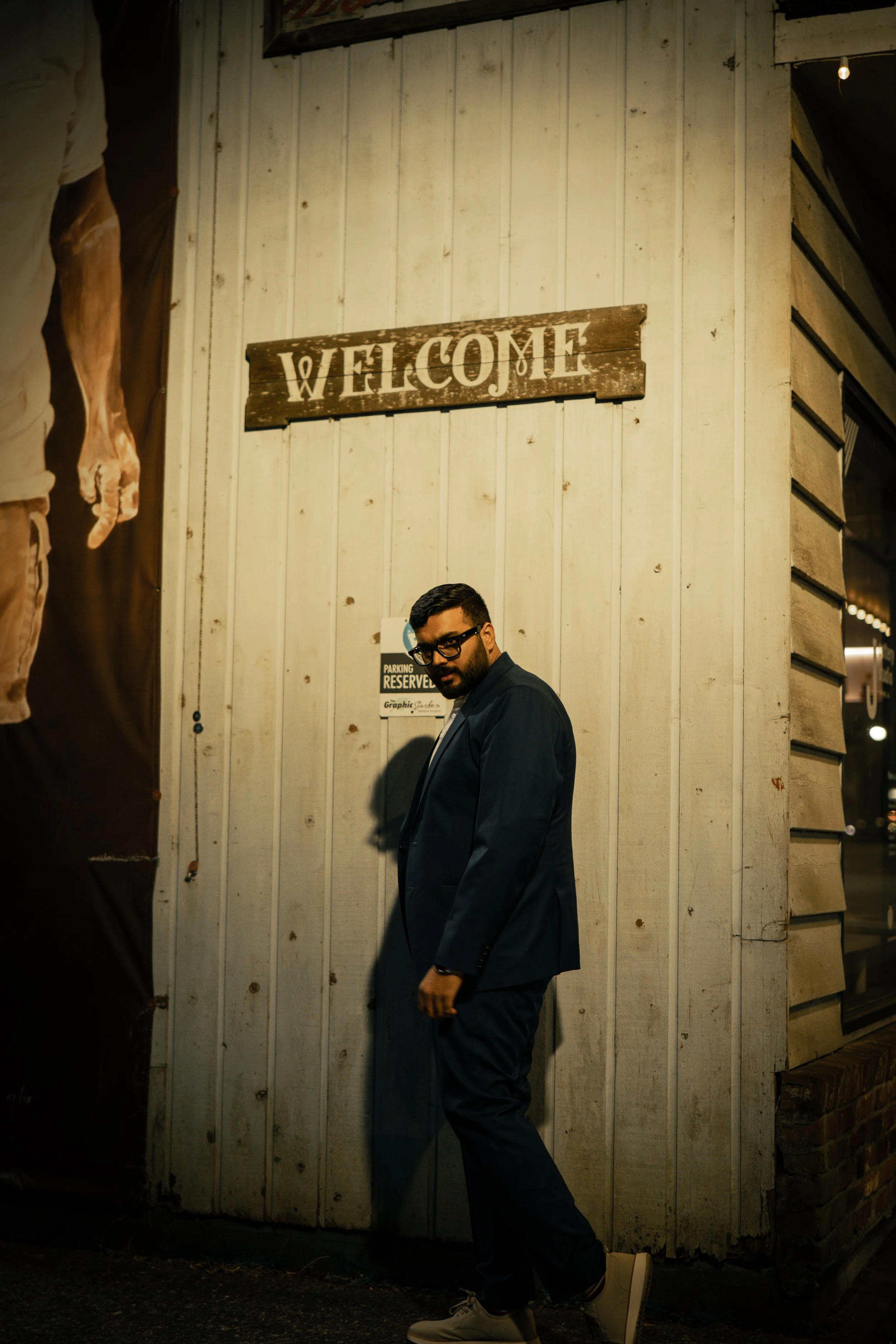 Man in a dark suit and glasses standing next to a white wooden wall with a "Welcome" sign above and a parking notice below, in a dimly lit setting.
