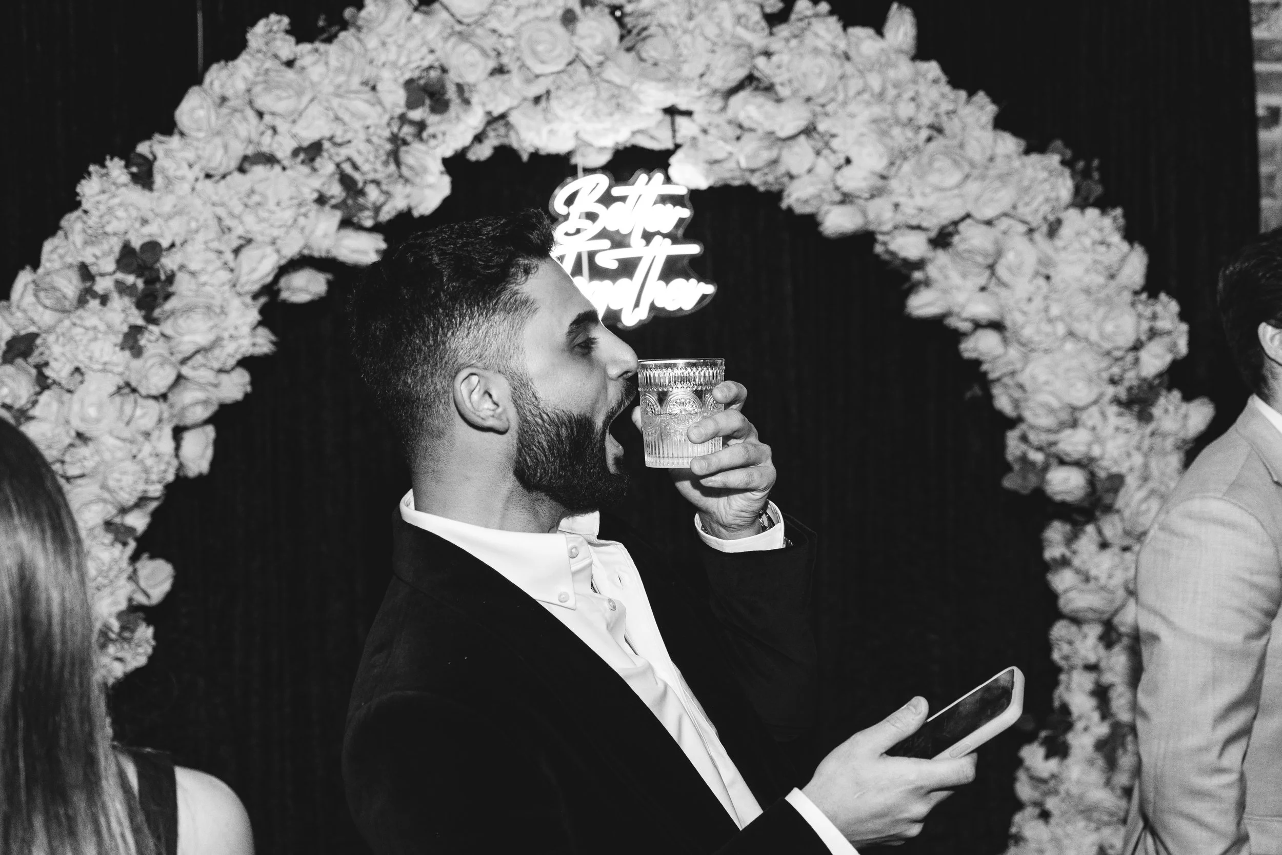Black and white photo of a man holding a drink and a phone, standing in front of a floral arch with a neon sign.