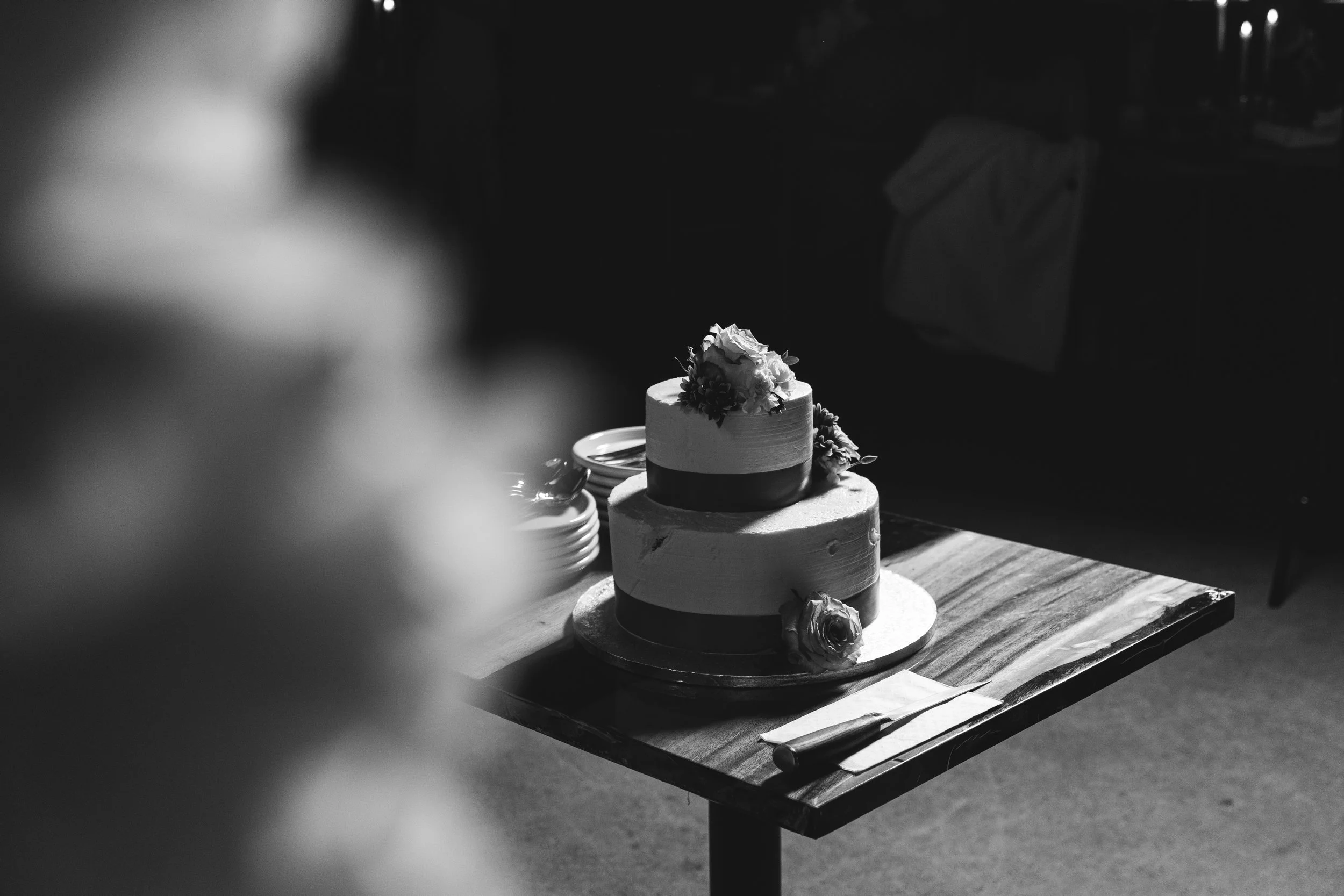 Black and white photo of a two-tiered cake on a wooden table with flowers on top, plates, and cutlery nearby.