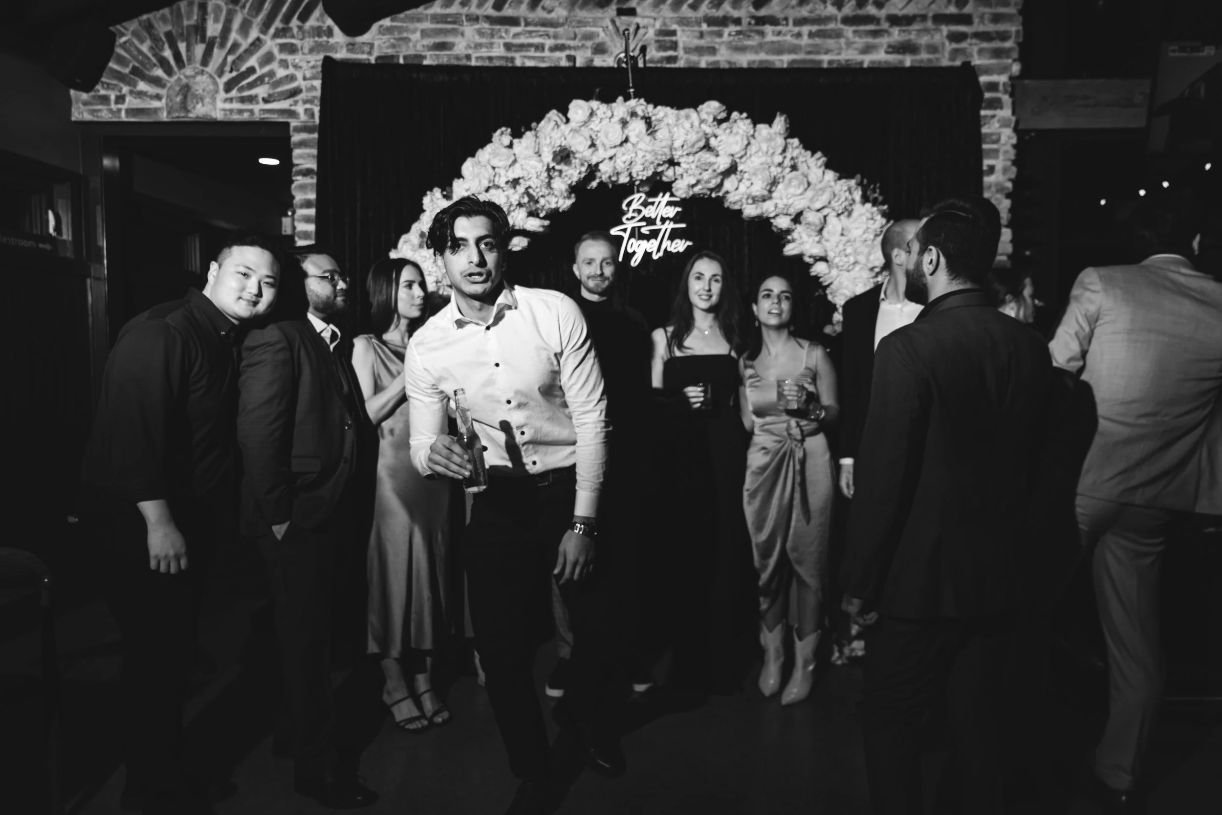 Group of people at a party in front of a flower arch with a 'Better Together' sign, in black and white.