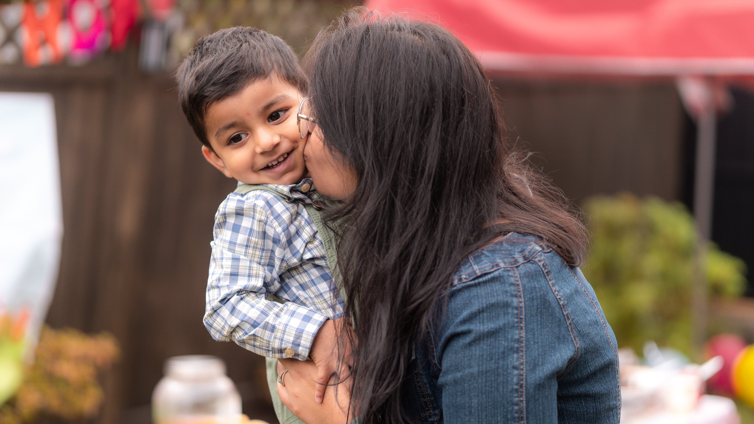 A woman with long dark hair and glasses kisses a smiling child in a plaid shirt on the cheek outdoors. The background is blurred, with colorful decorations visible.