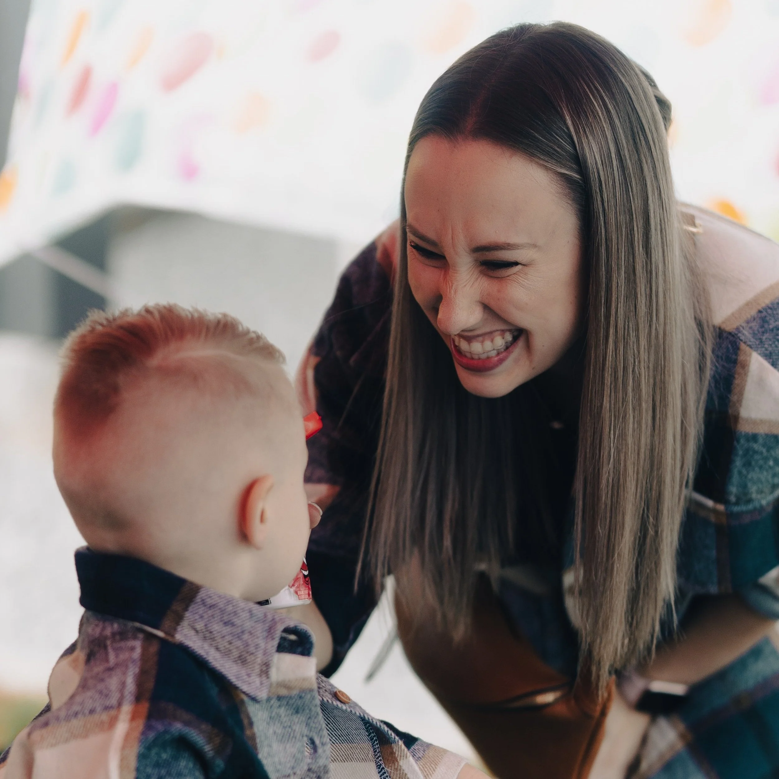 A woman with long hair smiling at a young child. Both are wearing plaid shirts. The background is out of focus, with colorful shapes visible.