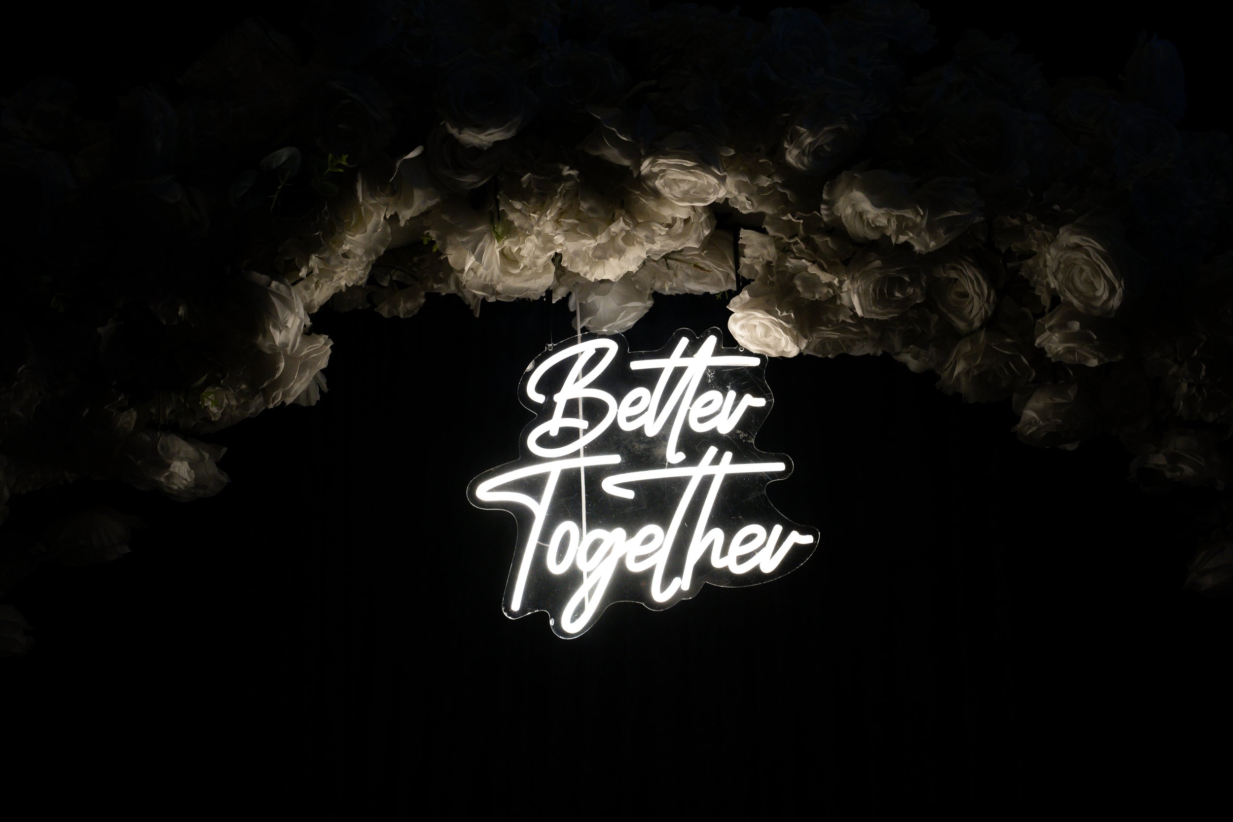 "Better Together" neon sign surrounded by white flowers on a dark background.