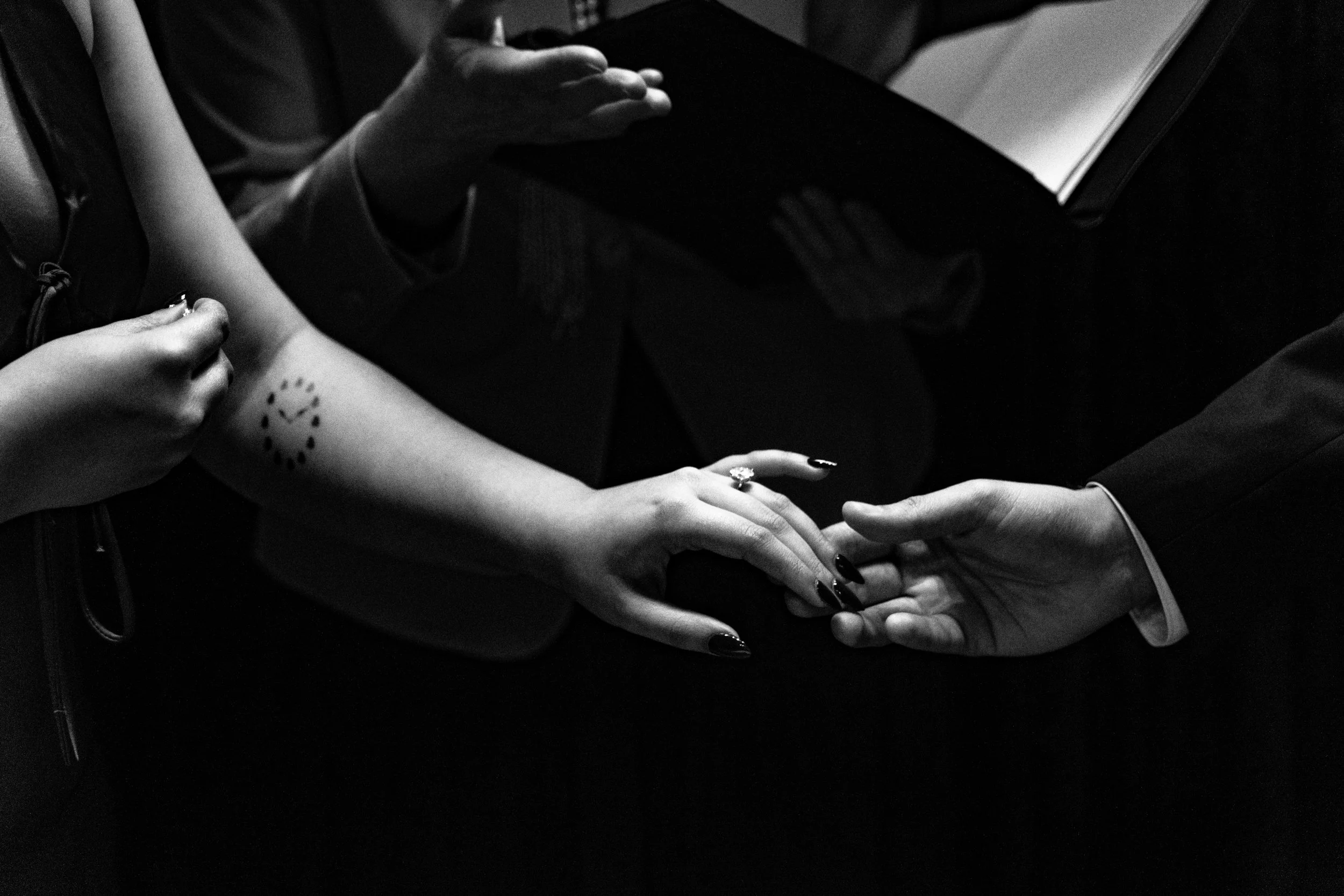 Black and white image of a couple holding hands during a ceremony. One person has a tattoo on their arm and the other wears a suit. A person officiating holds a book.