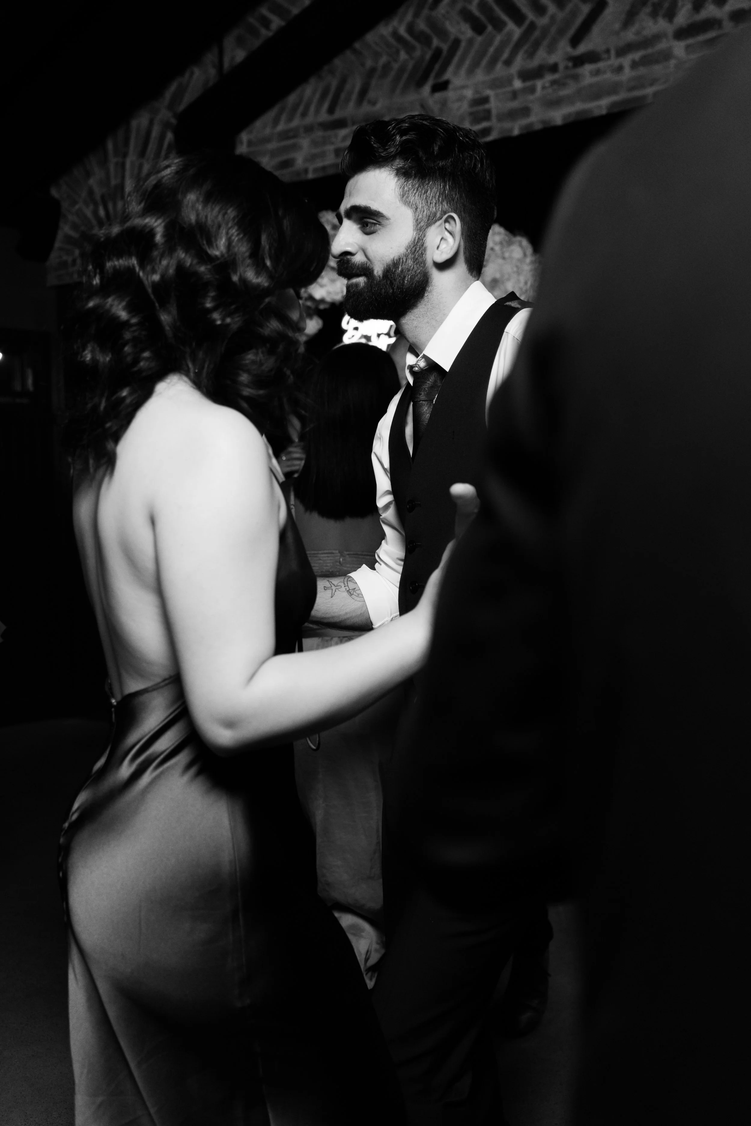 Black and white photo of a couple dancing closely, with the woman in a backless dress and the man in a suit, in a dimly lit room.